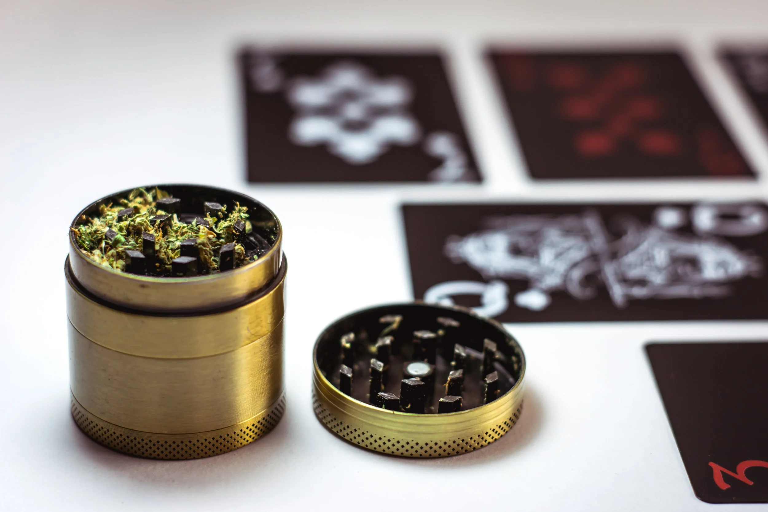 Two metal grinder containers with cannabis inside and playing cards on a white surface