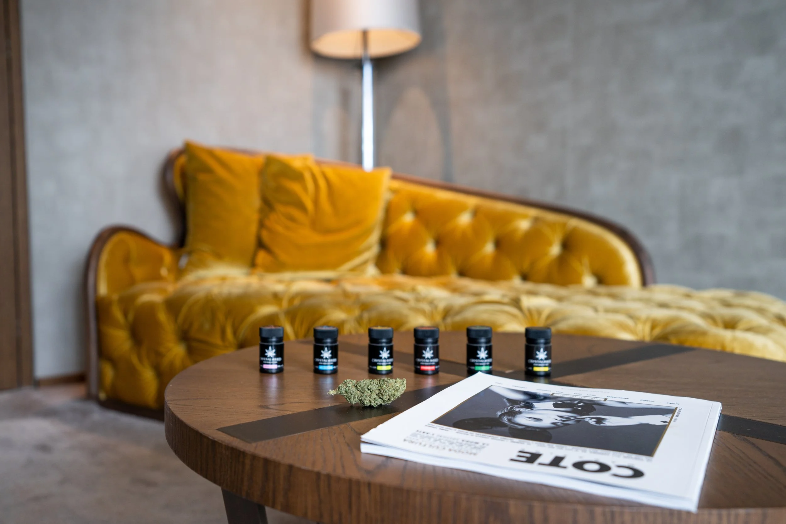 A wooden table with five small bottles of cannabis oil, a cannabis bud, and a black-and-white magazine on top, in a room with a yellow velvet tufted sofa and a floor lamp in the background.