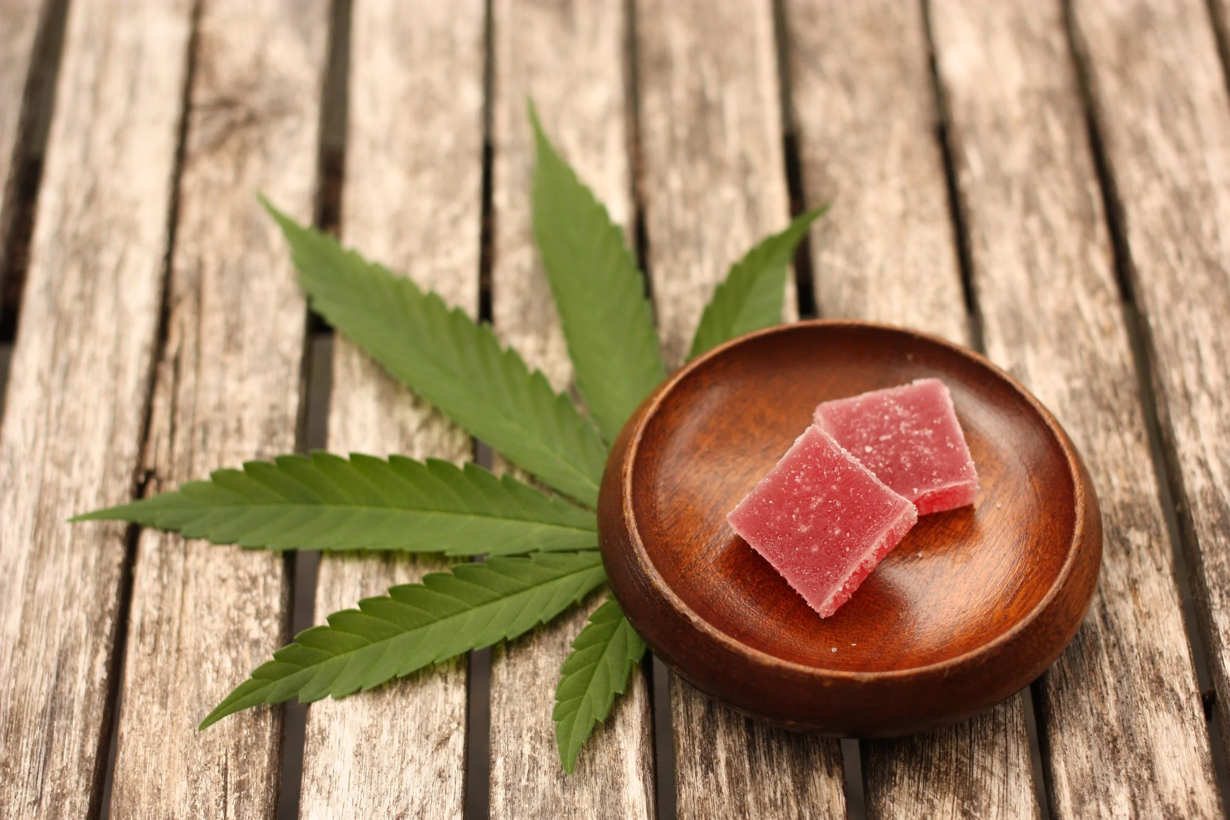 Two pink THC-infused chocolate squares on a wooden dish with cannabis leaves underneath, on a weathered wooden surface.