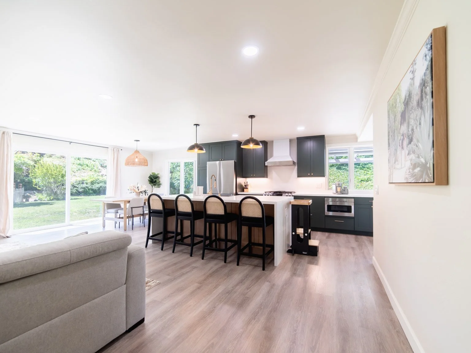 Open-concept living area with a kitchen featuring black cabinets, a white island with four black bar stools, a dining table with white chairs, and large windows showing a green backyard.