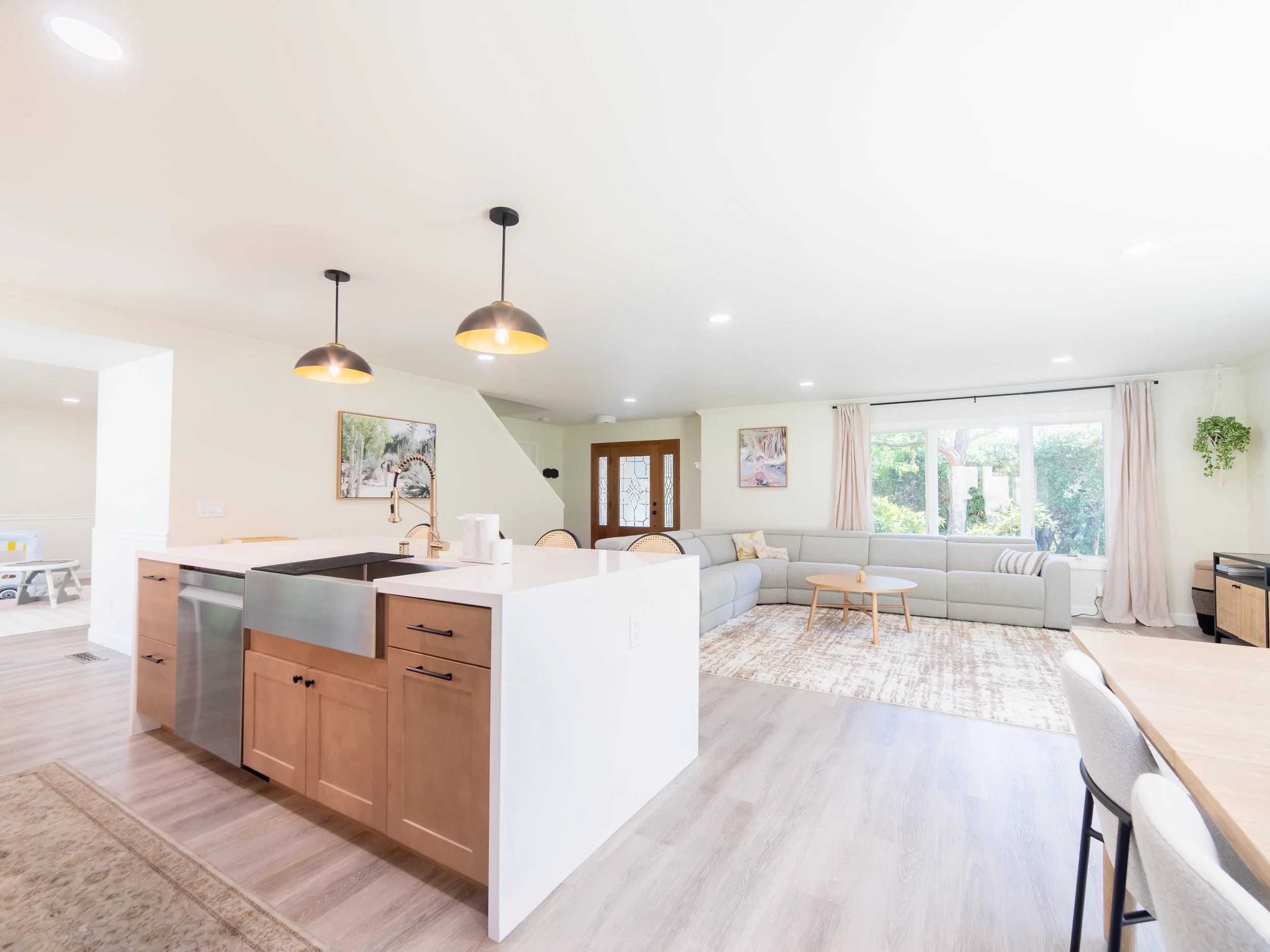 Bright living room with large window, beige sectional sofa, wooden coffee table, area rug, and open kitchen with white island and wooden cabinets.
