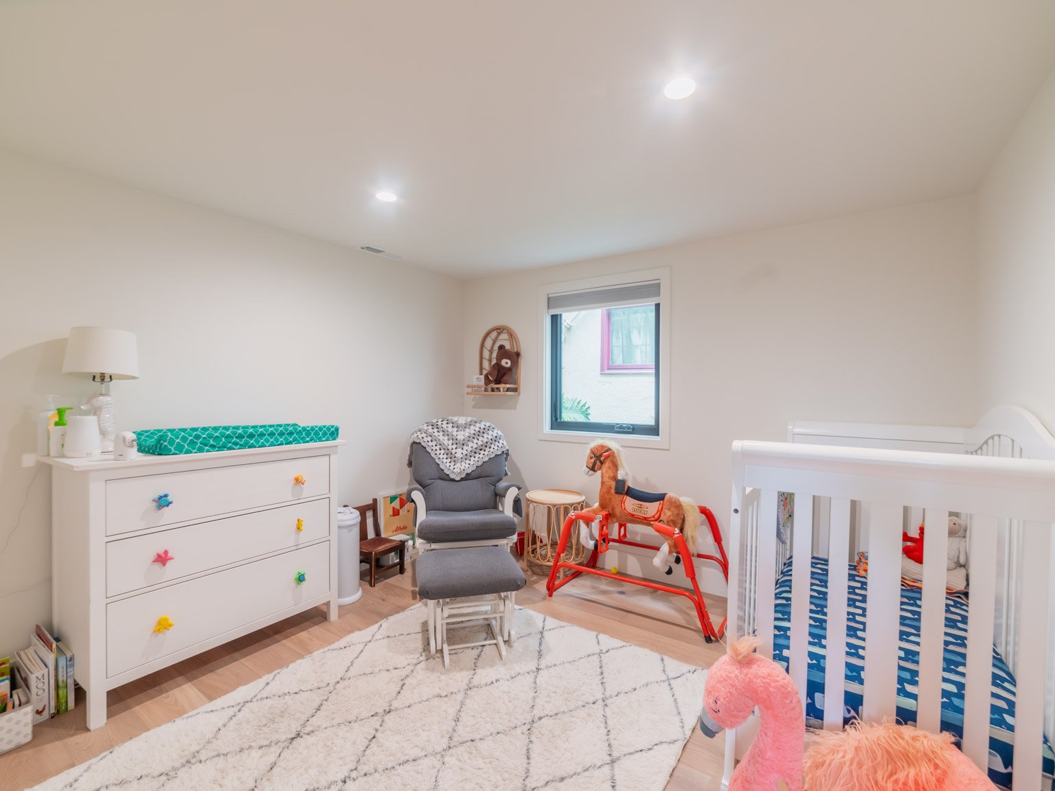 Child's nursery with white crib, plush pink flamingo, rocking horse, gray chair, changing table with colorful knobs, window, bookshelf, and stuffed animal toys.