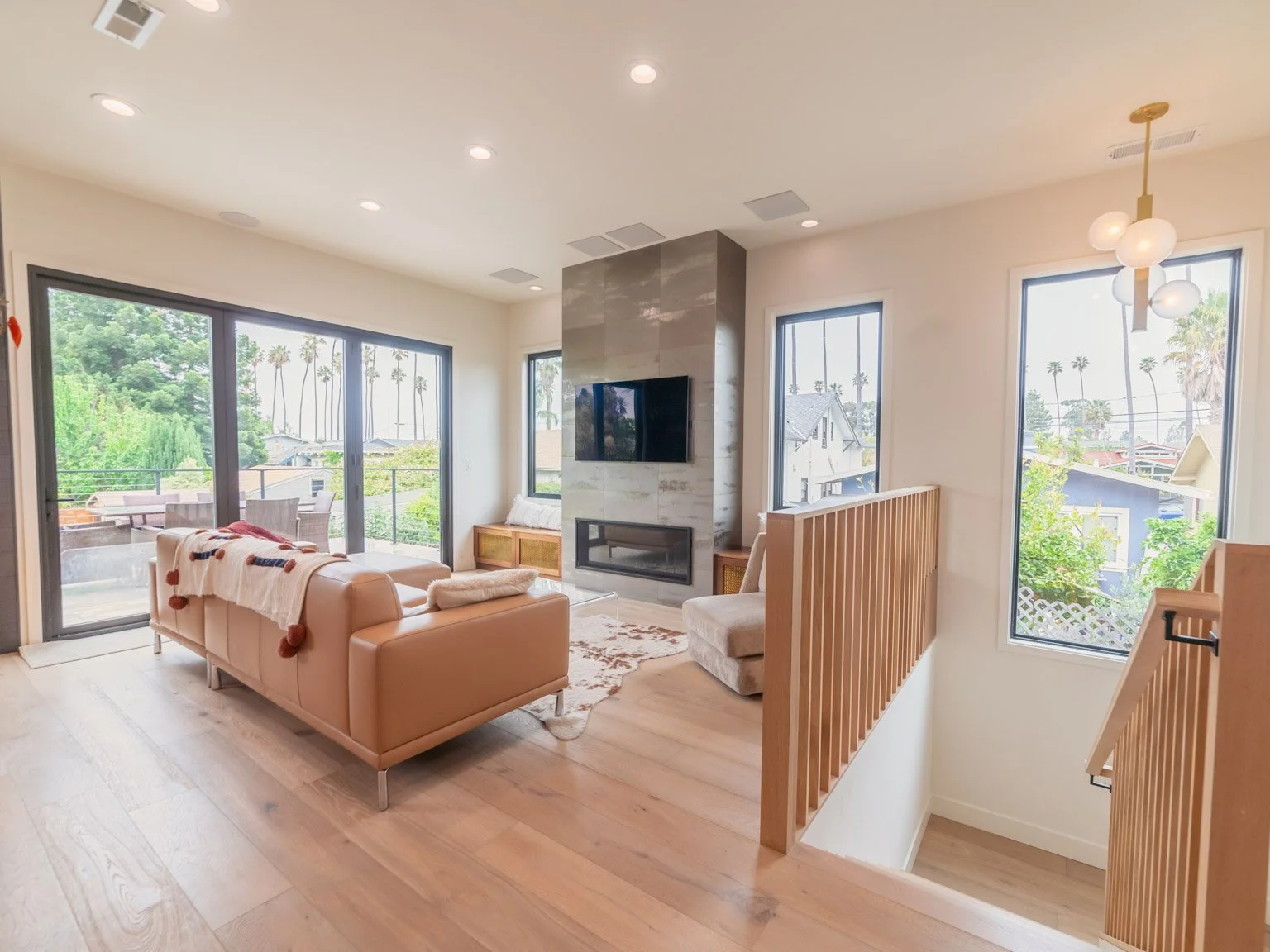 Living room with large windows showing palm trees, beige leather sofa, fireplace with mounted TV, wooden staircase railing, and a modern chandelier.