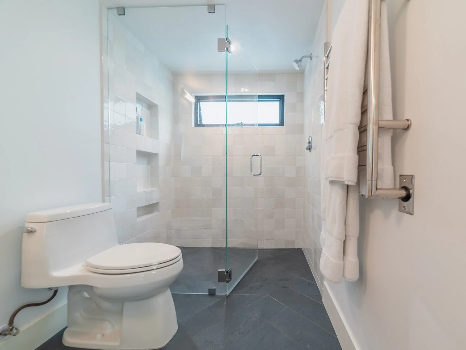 Modern bathroom with a toilet on the left, a walk-in glass shower in the center, and a towel radiator on the right side with white towels hanging.