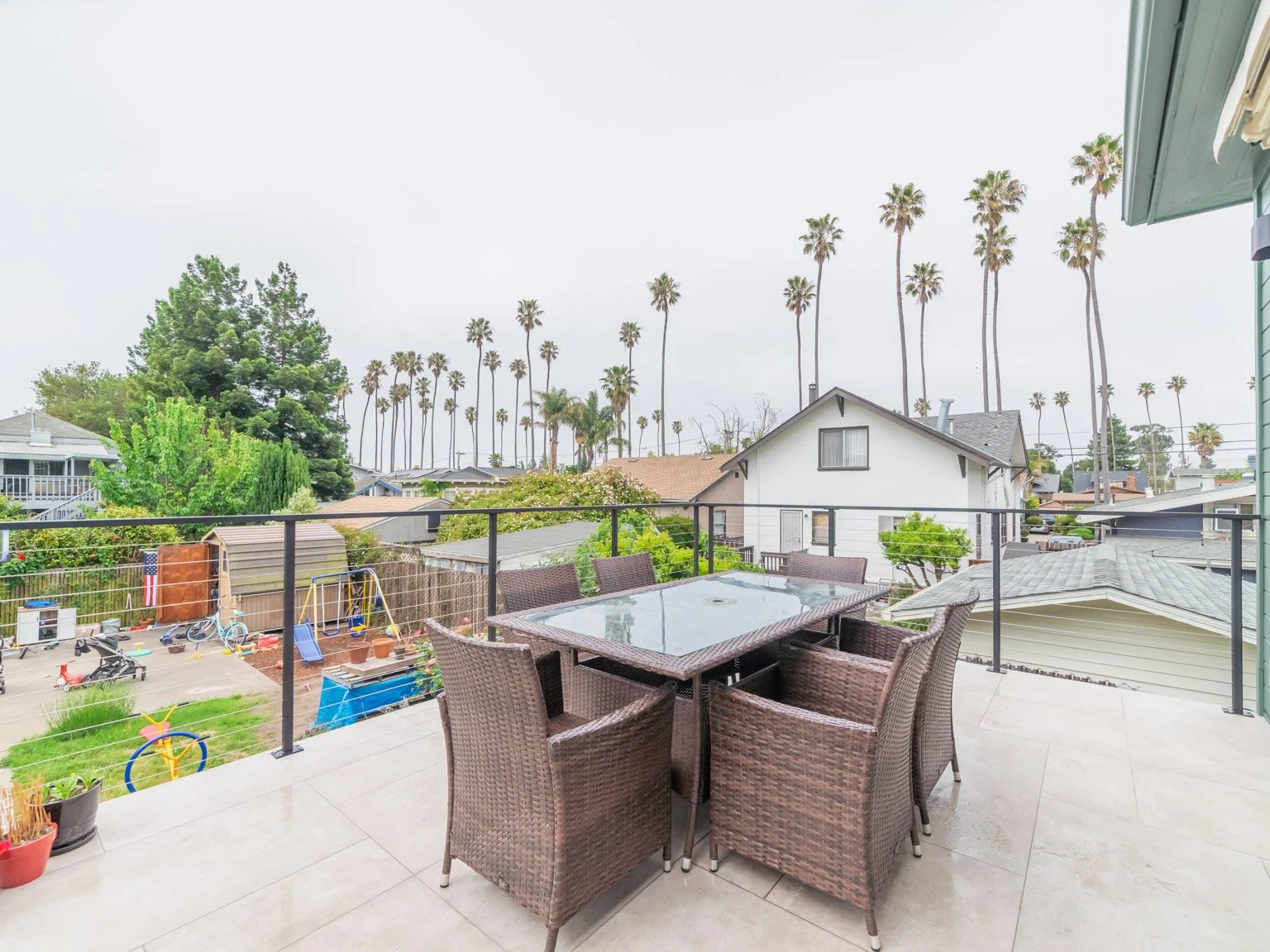 This is a balcony with a glass-top dining table and six wicker chairs, overlooking a backyard with children’s play area and a neighborhood with houses and tall palm trees in the background.