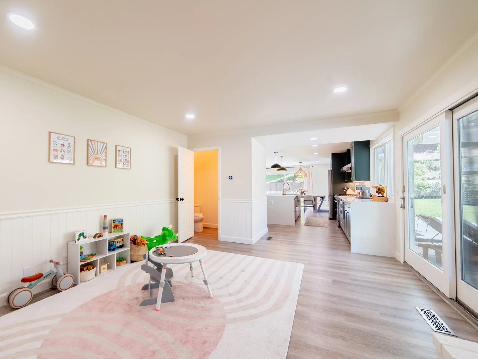 A bright, open-concept living space with a children's play area, a small table, a toy storage unit, and a view into the kitchen and dining area with large windows and sliding doors.