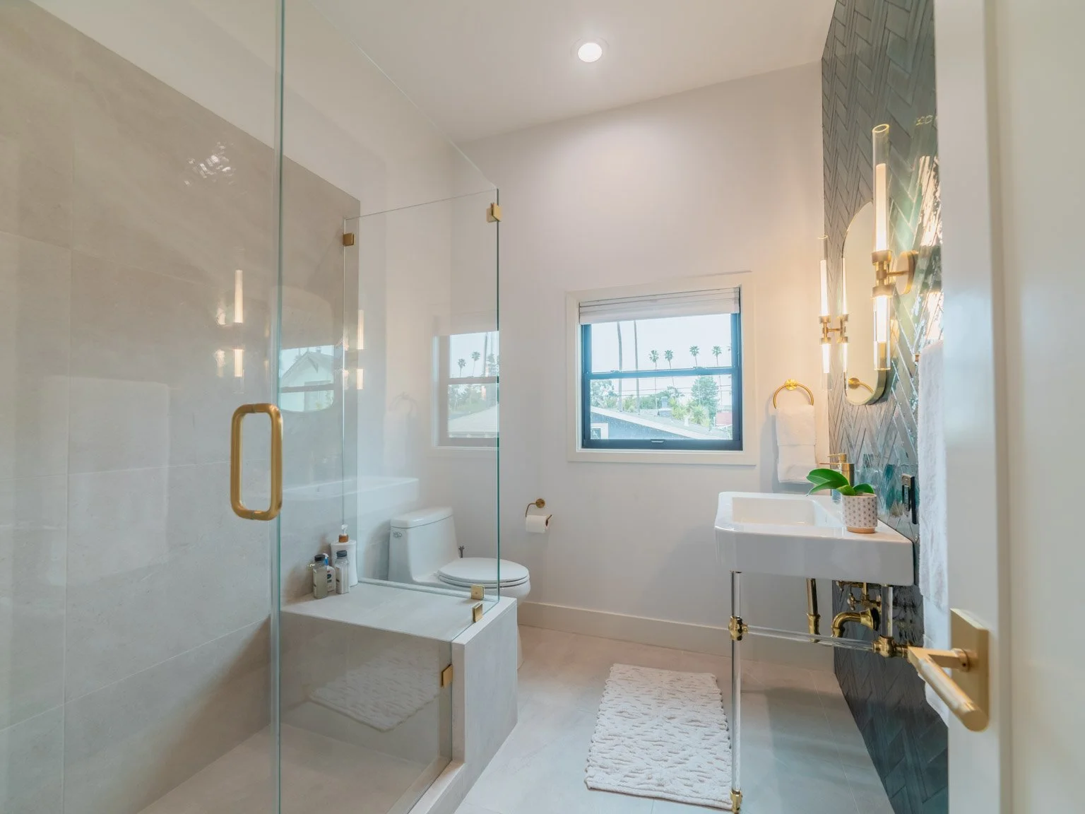 Modern bathroom with a glass shower enclosure, white toilet, white sink with gold fixtures, and a window with a view of palm trees.