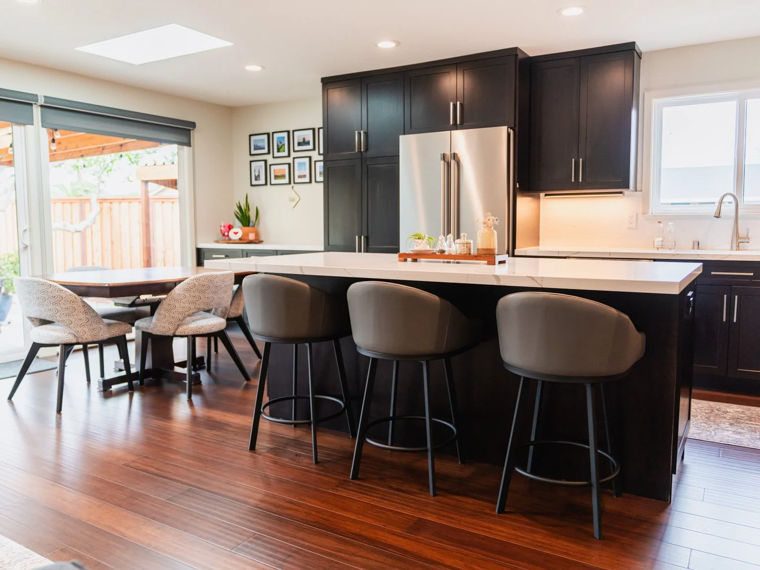 Modern kitchen with black cabinets, stainless steel refrigerator, white countertop, island with three beige barstools, wood flooring, sliding glass door, and picture window.