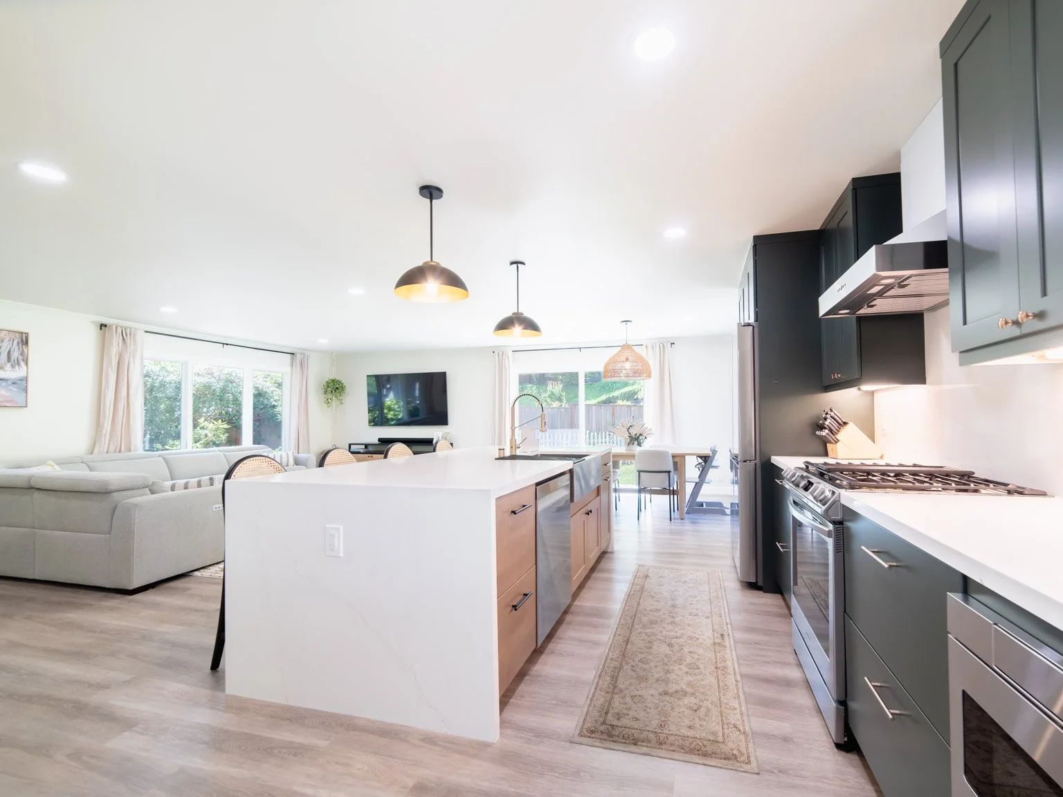 Open-concept kitchen and living room with white island, stainless steel appliances, black cabinets, beige dining chairs, large windows, television, and minimal decor.
