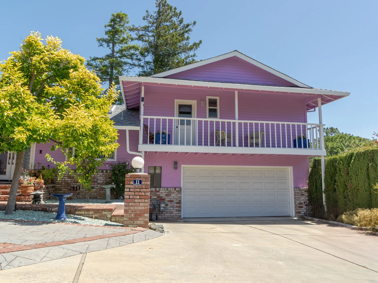 A two-story house painted in purple with a white garage door, a balcony with white railings, and surrounded by trees and shrubs on a sunny day.