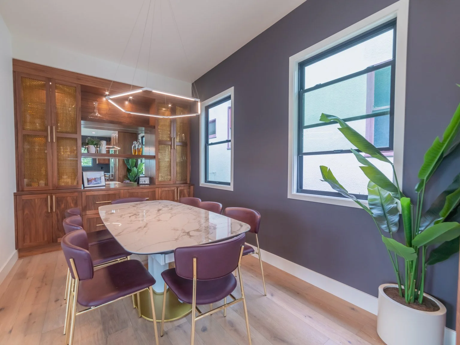 Modern dining room with a marble-top table, purple chairs, wooden built-in storage, large windows, and a potted plant.