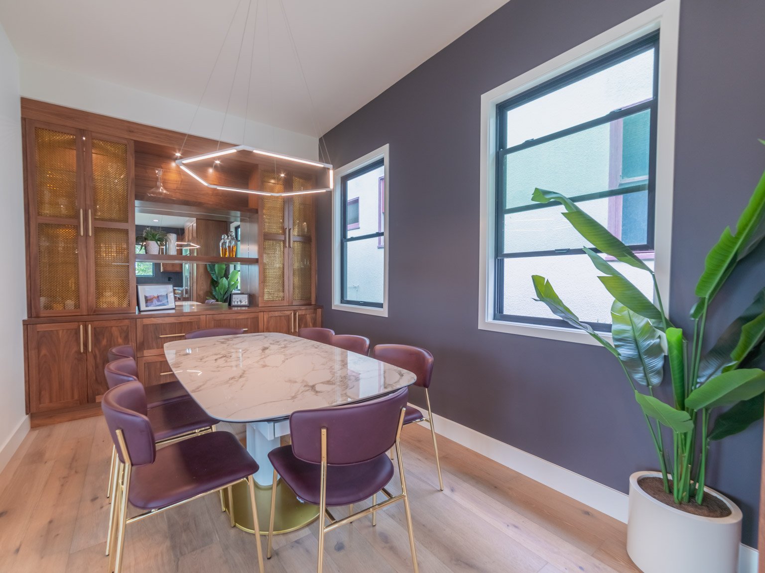 Modern dining room with a marble-top table, purple chairs, wooden built-in storage, large windows, and a potted plant.