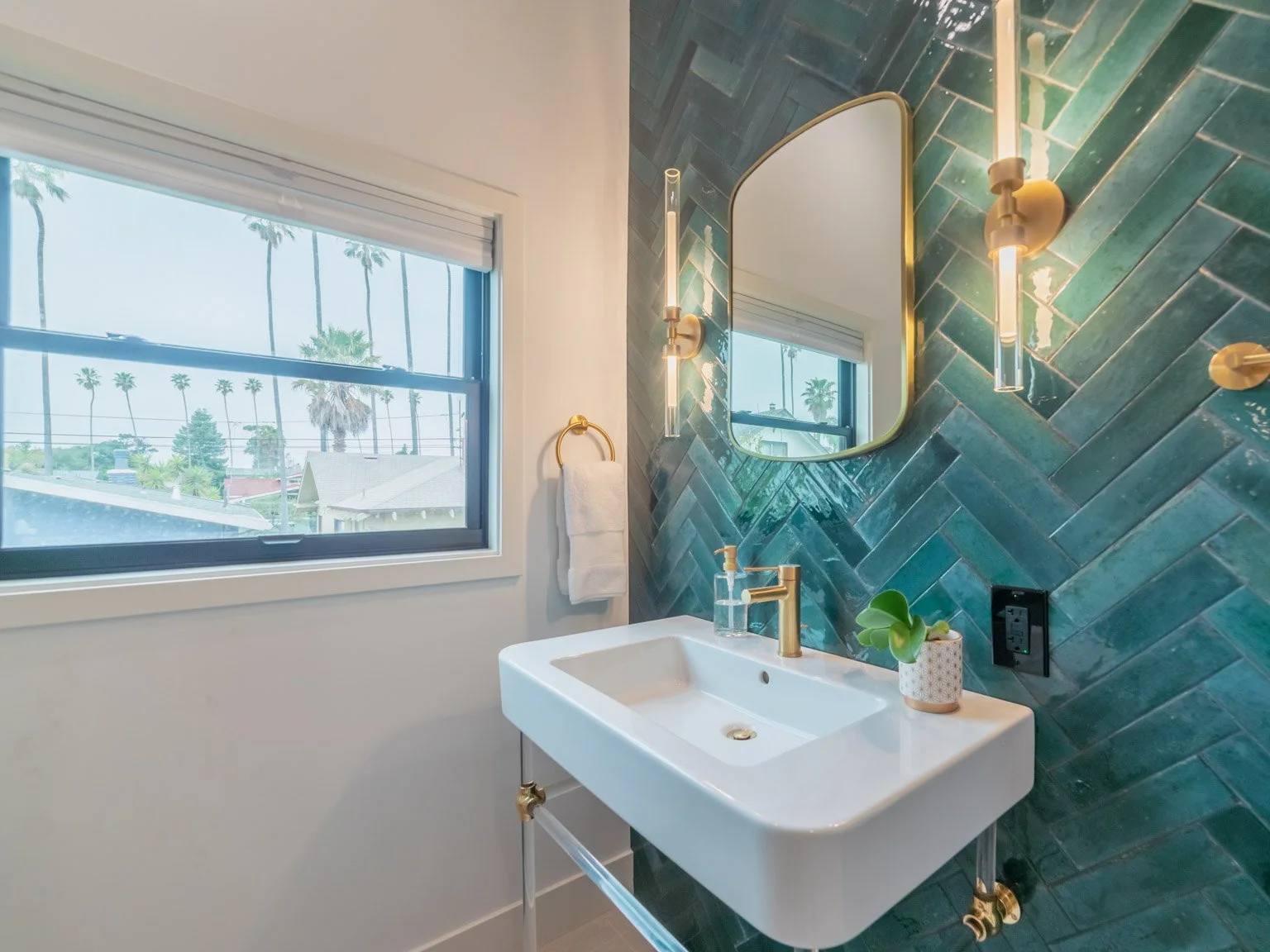 Bathroom with a white sink, gold fixtures, a decorative mirror, a wall-mounted light, a window showing palm trees outside, a towel ring with a white towel, a small plant on the sink, and a teal-tiled accent wall.