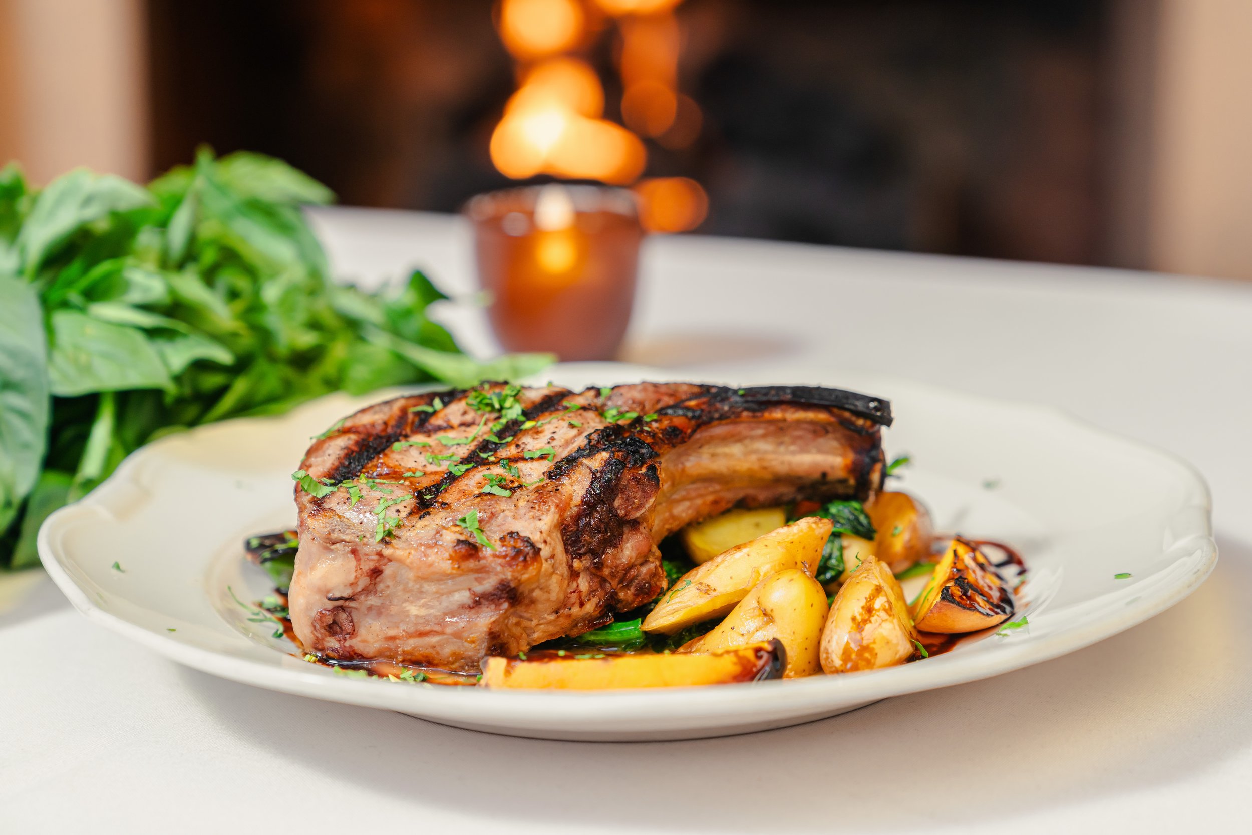 A grilled pork chop garnished with chopped herbs served with roasted garlic and vegetables on a white plate