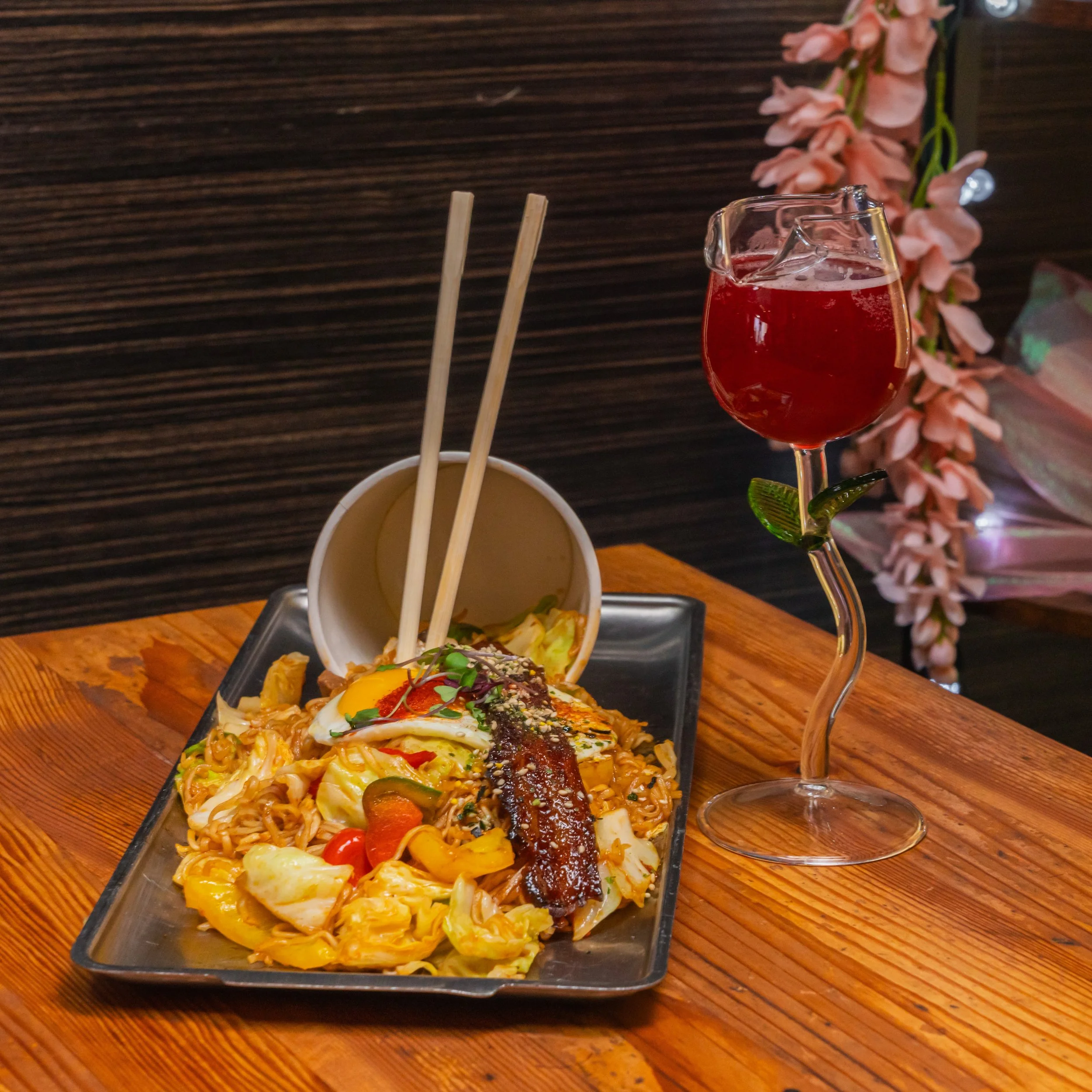 Plate of Asian stir-fried noodles with vegetables, served with a sliced boiled egg and garnished with sesame seeds, accompanied by a cup of soup and a glass of red cocktail on a wooden table.
