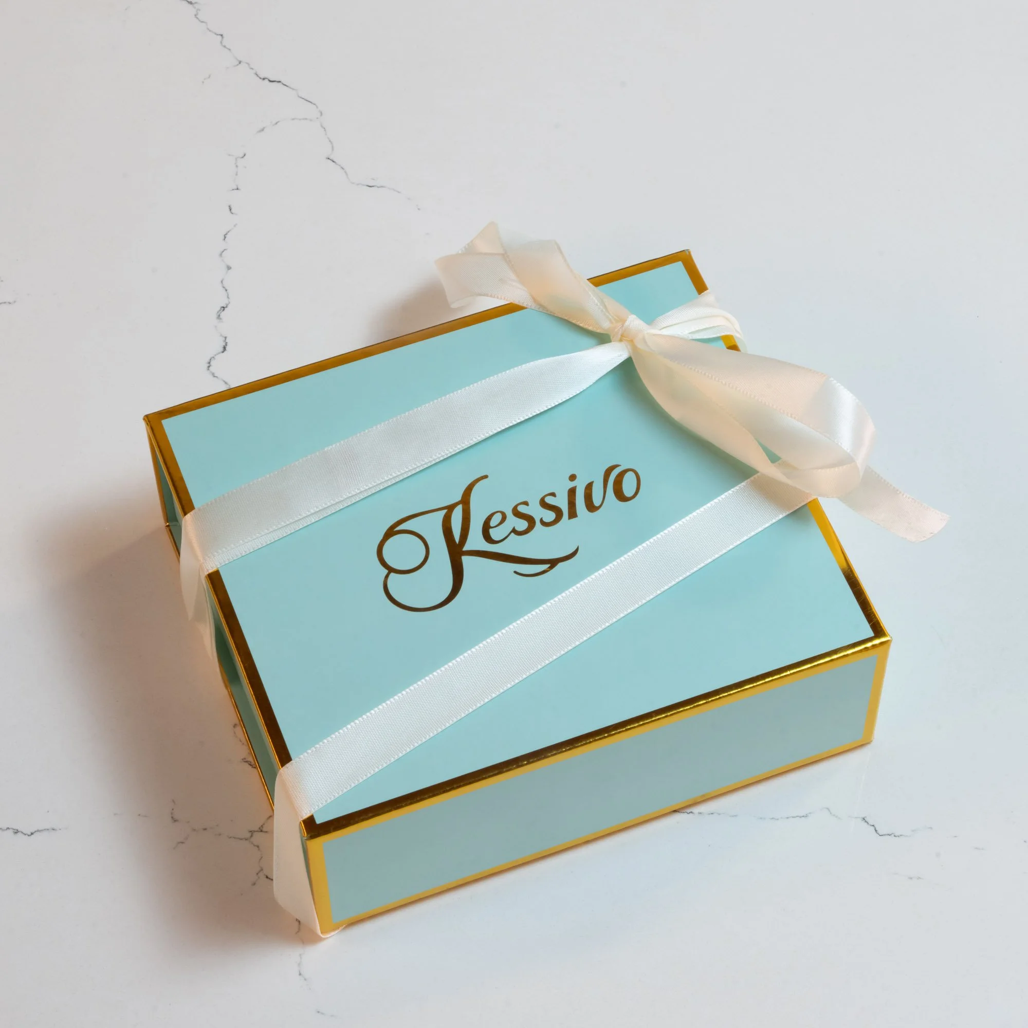A light blue gift box with a white satin ribbon and bow, featuring the name 'Kessio' written on the top, placed on a white surface with faint cracks.