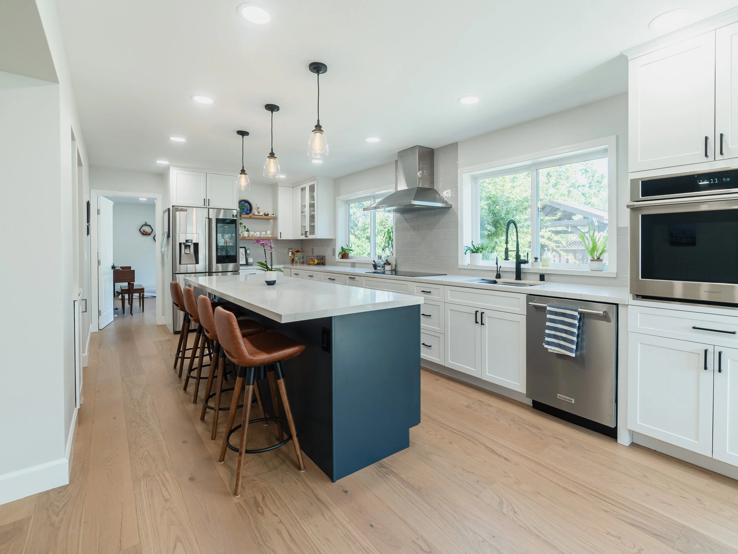 Modern white kitchen with an island, pendant lighting, stainless steel appliances, and large windows with green outdoor views.