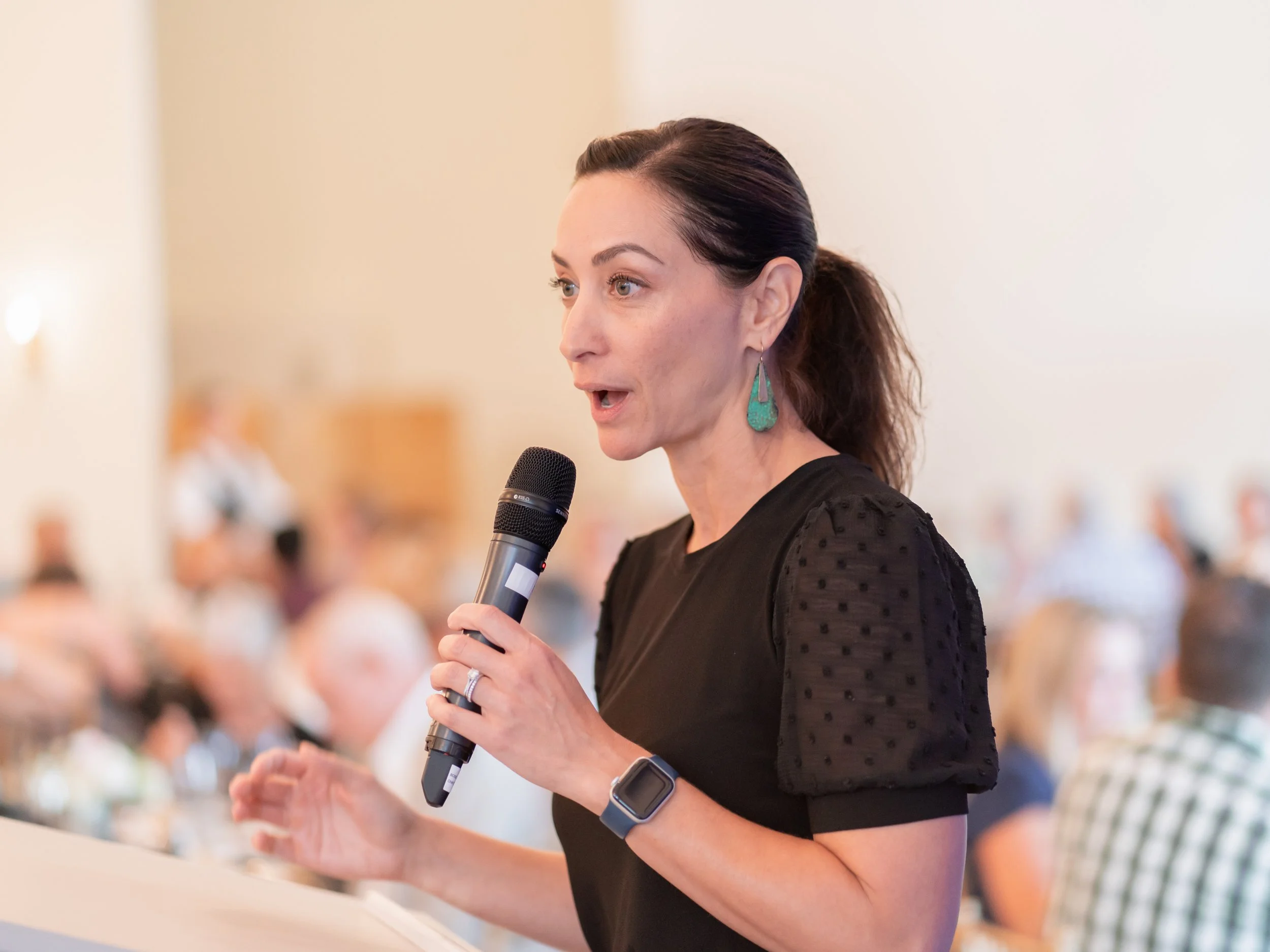 A woman holding a microphone and speaking at a conference or event with an attentive audience in the background.