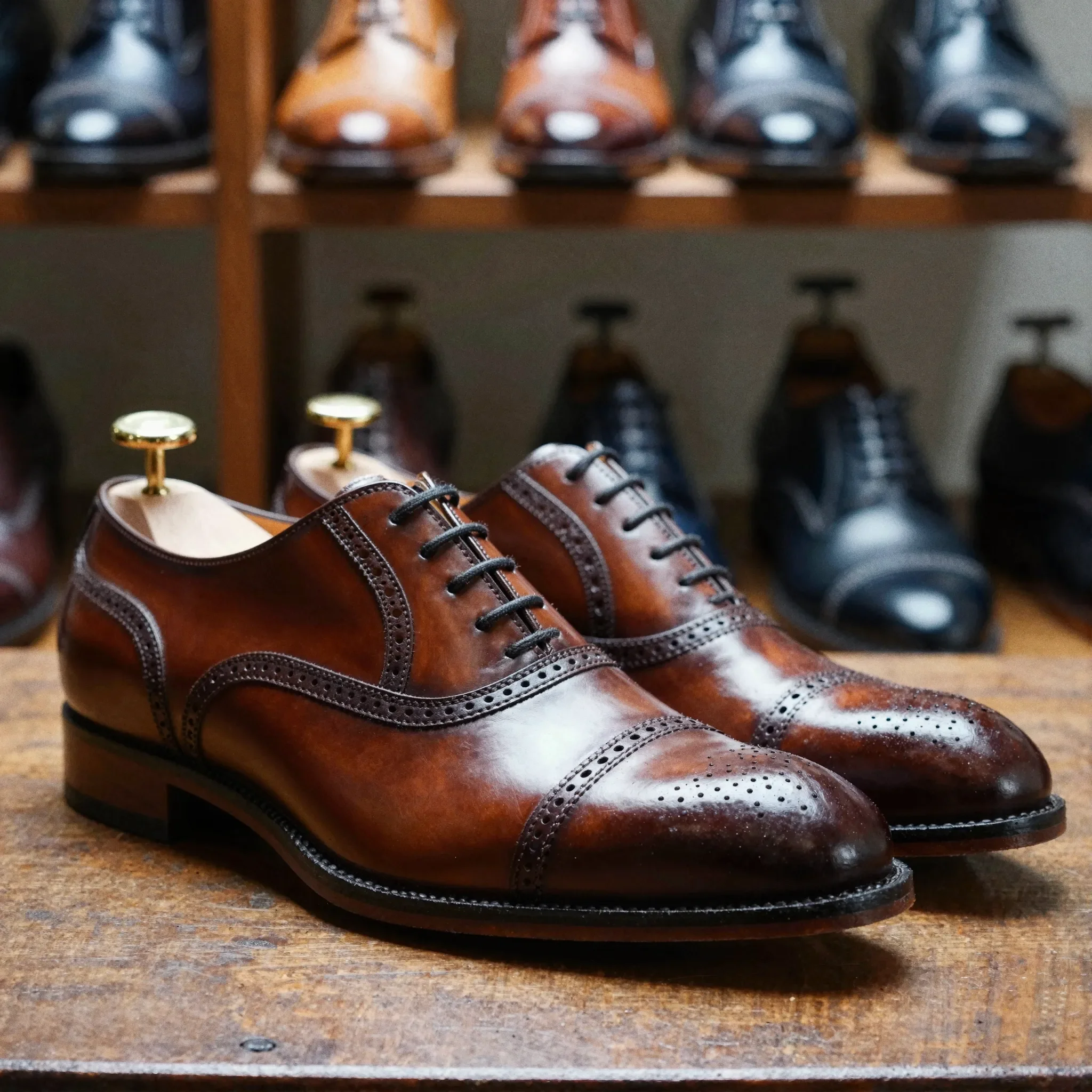 A pair of men’s leather oxford dress shoes with brogue detailing on a wooden surface, with additional shoes displayed on shelves in the background.