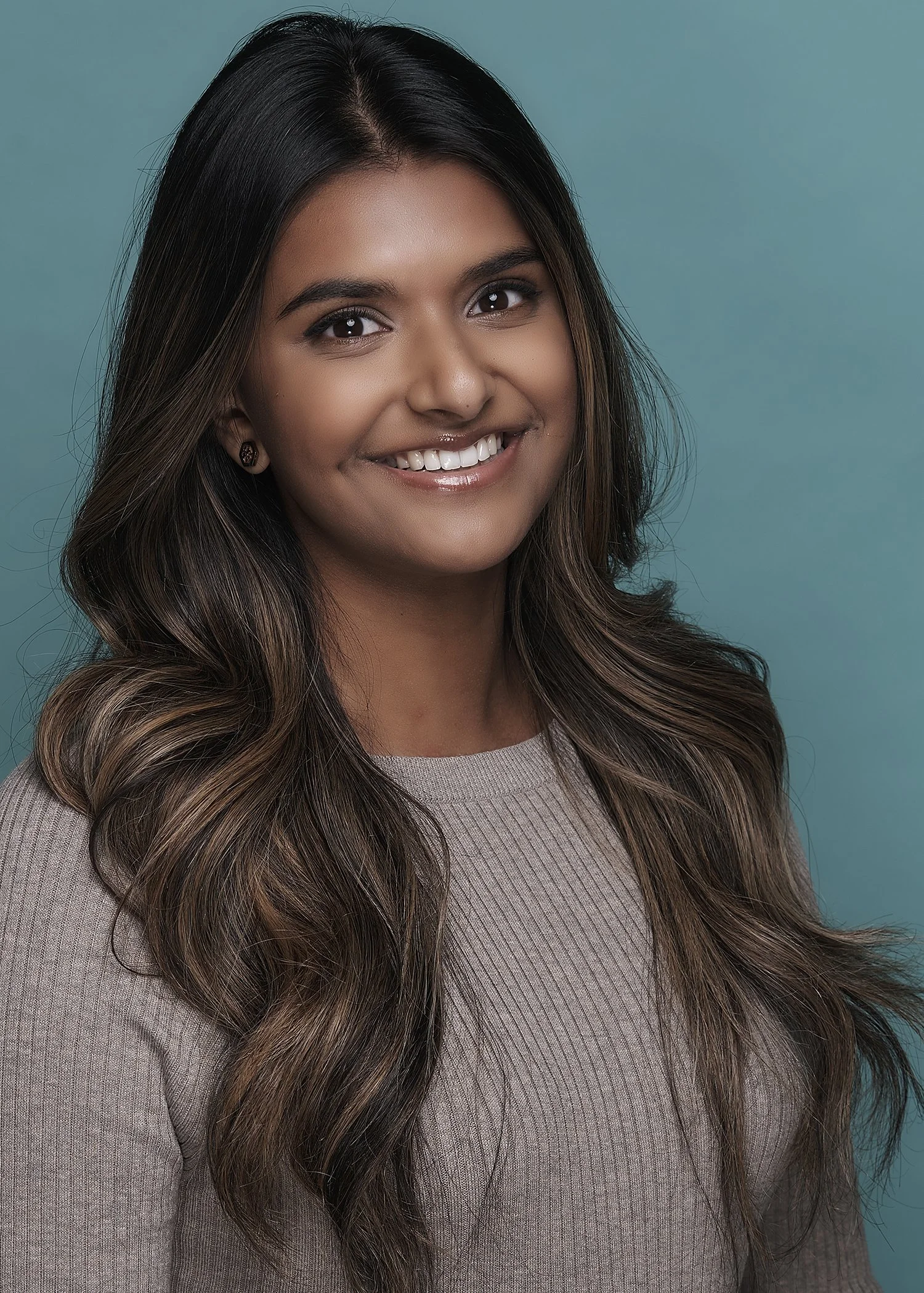 A young woman with long, wavy brown hair smiling at the camera against a teal background, wearing a beige top.
