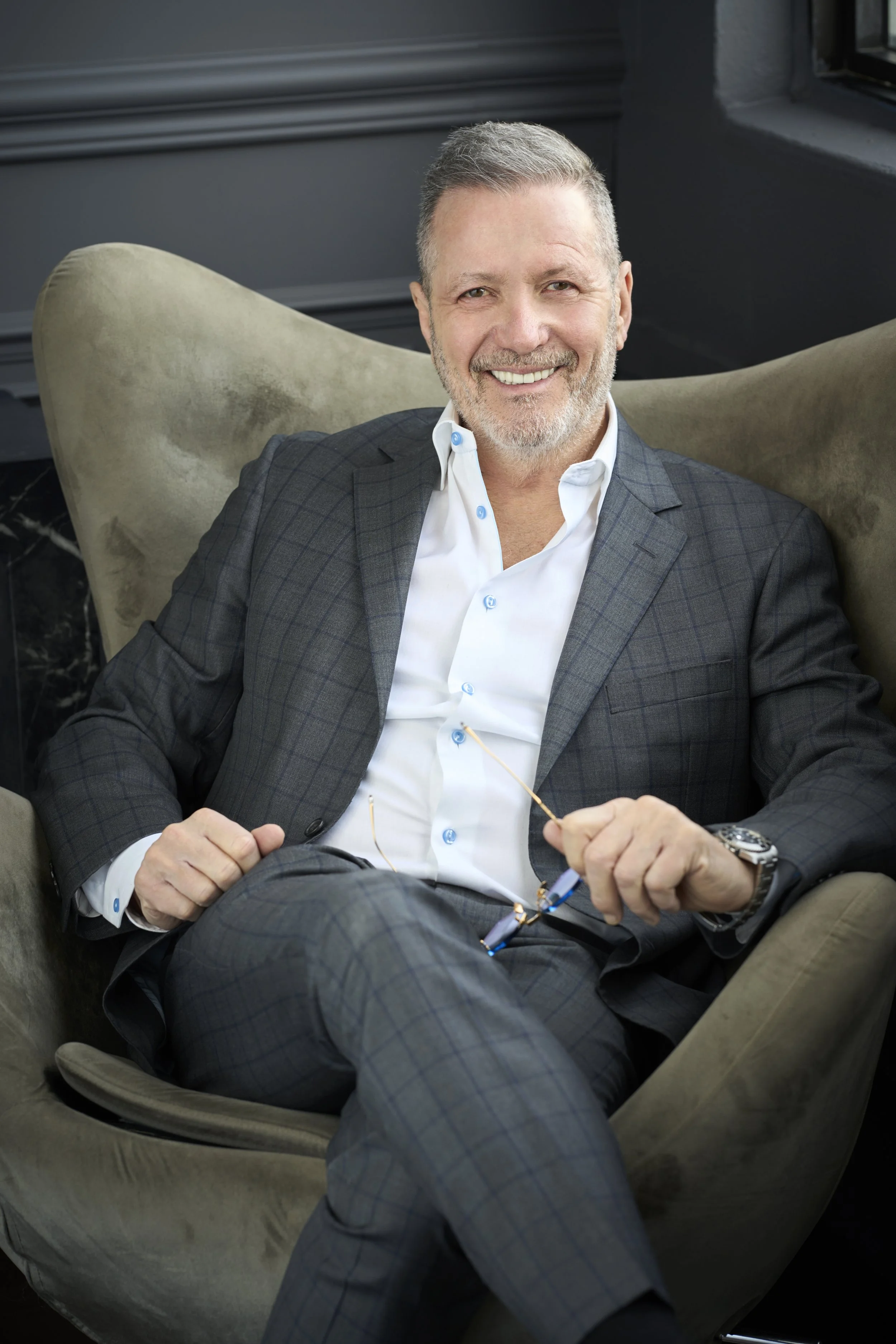 A smiling middle-aged man with gray hair and a beard, wearing a dark checkered suit and white shirt, sitting relaxed in a beige velvet armchair with crossed legs, holding glasses in his hand.