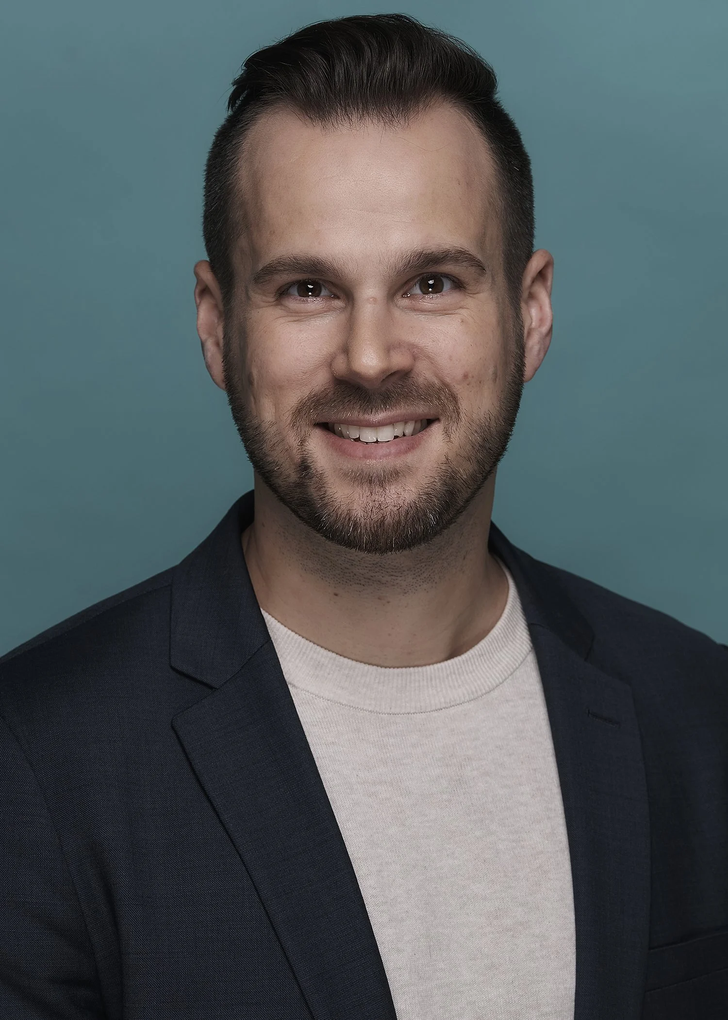 Headshot of a smiling man with short dark hair, a beard, wearing a dark blazer over a light-colored shirt, against a teal background.