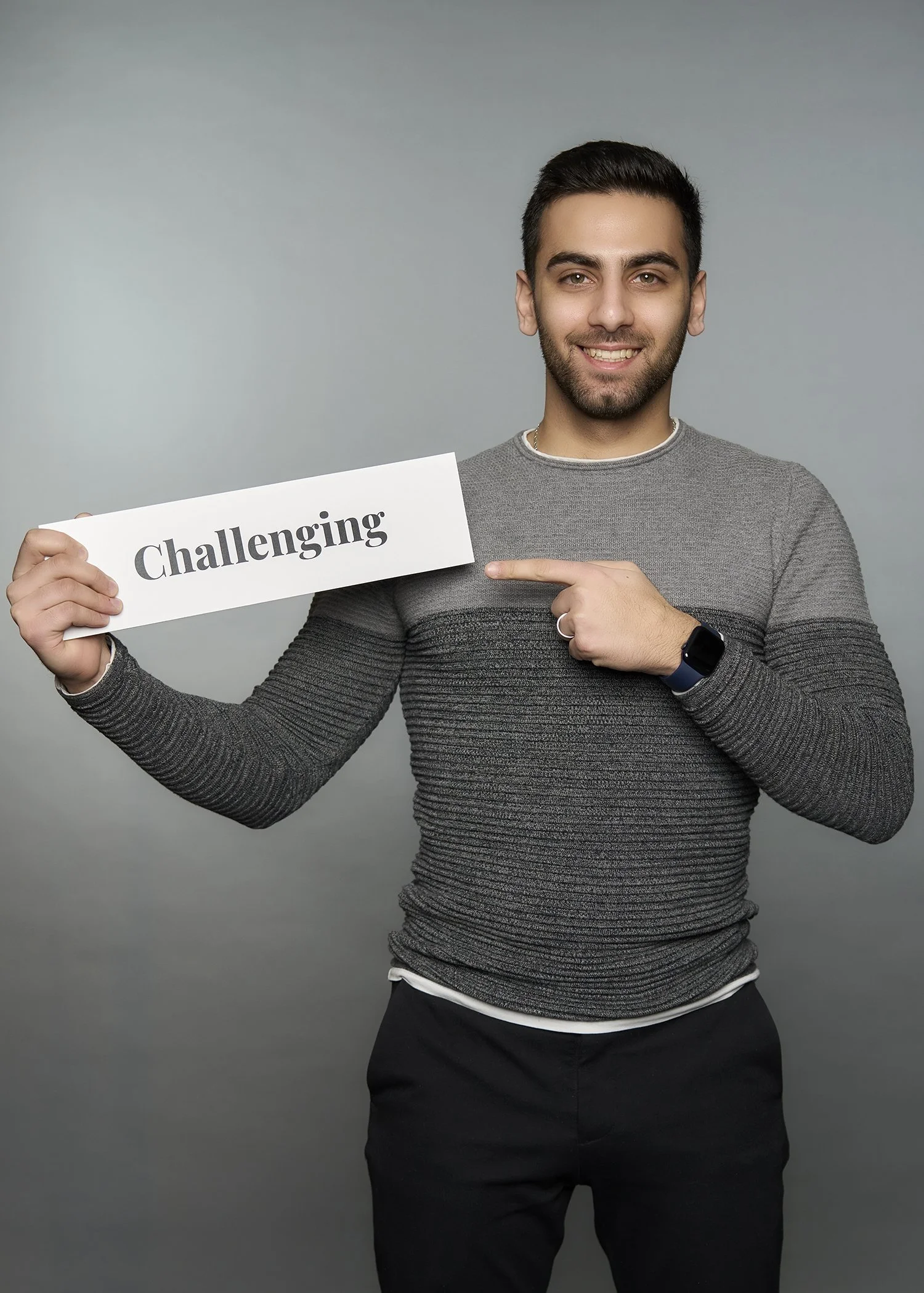 A young man with dark hair and a beard smiling, wearing a gray striped sweater and a smartwatch, holding a sign that says "Challenging" and pointing to it.