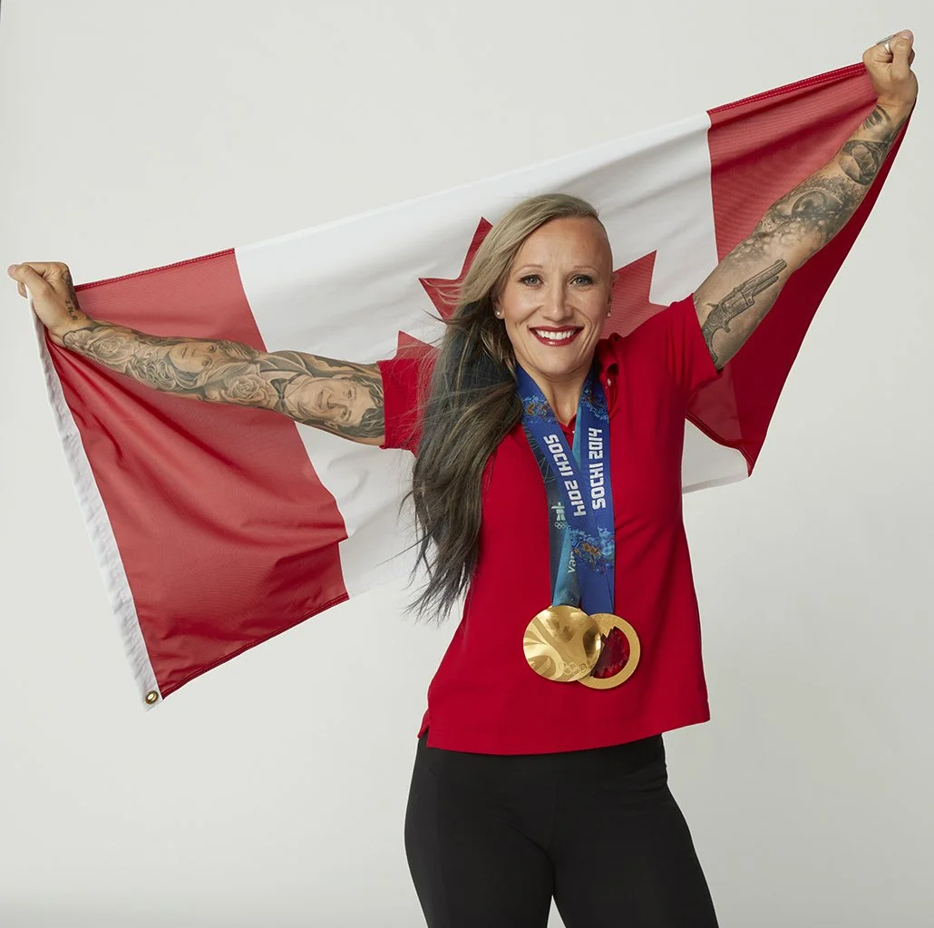 A woman with long hair and tattoos celebrating with a Canadian flag and wearing a gold Olympic medal, smiling against a plain background.