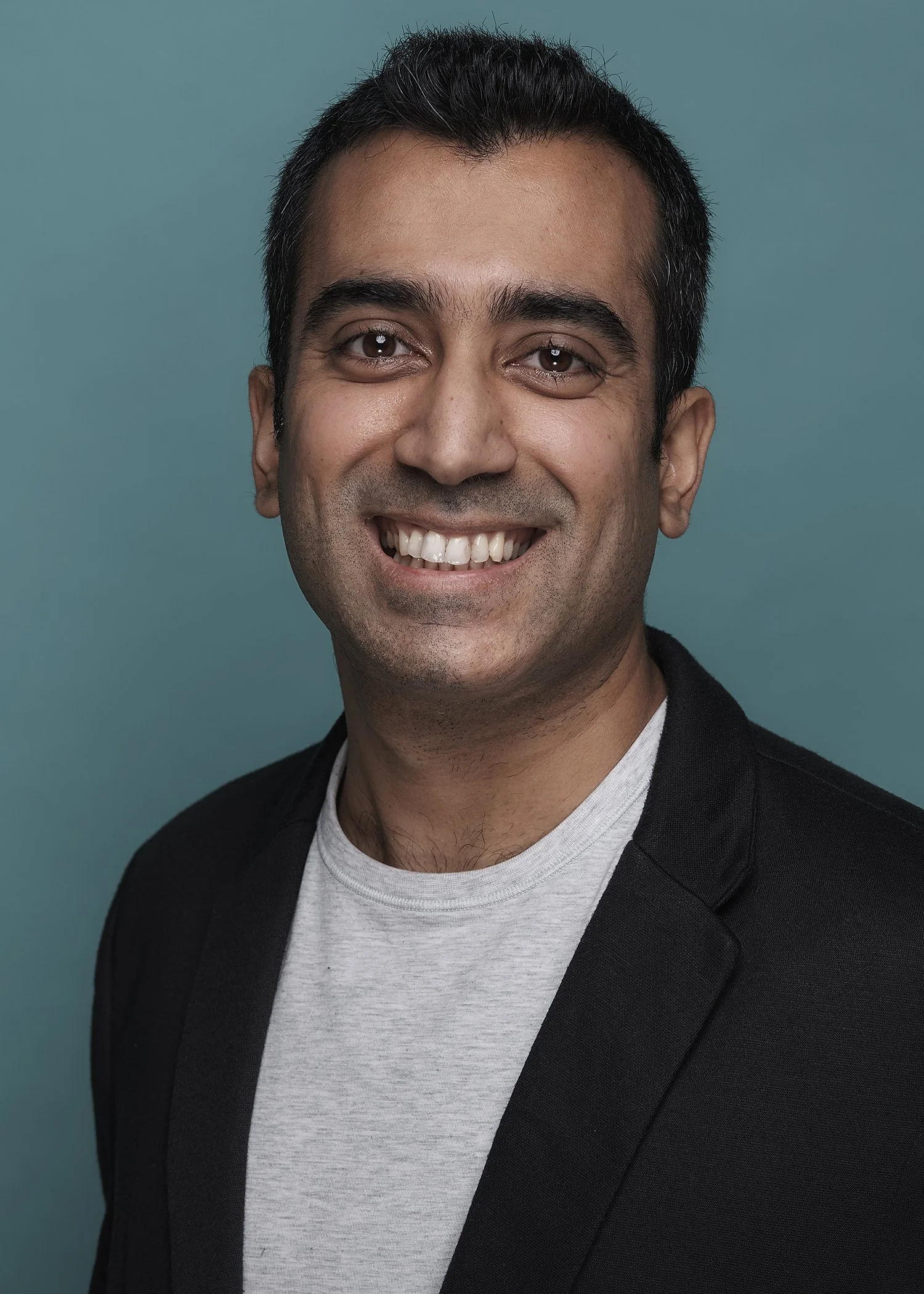 A smiling man with short dark hair wearing a black blazer over a light gray t-shirt against a teal background.