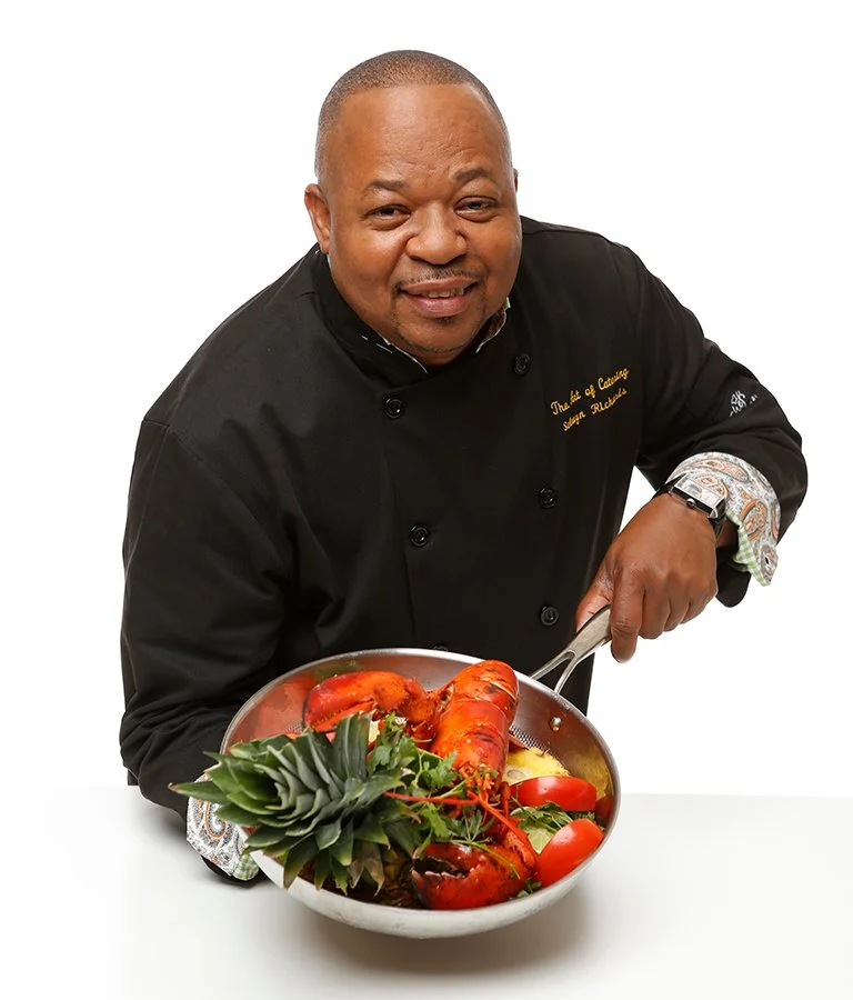 A chef in a black uniform holding a frying pan with lobster, pineapple, and tomatoes on a white background.