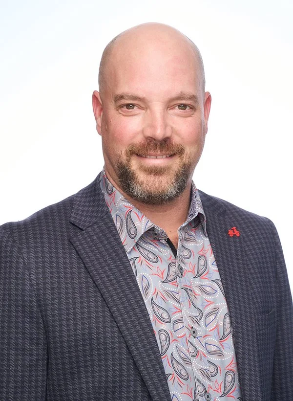A middle-aged man with a bald head and beard, smiling, wearing a patterned button-up shirt and a dark blazer with a small red pin on the lapel, against a plain white background.