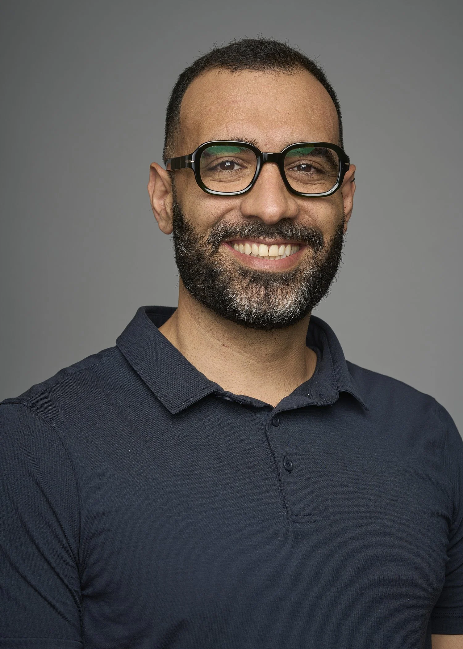A man with glasses and a beard smiling in a navy blue collared shirt against a gray background.