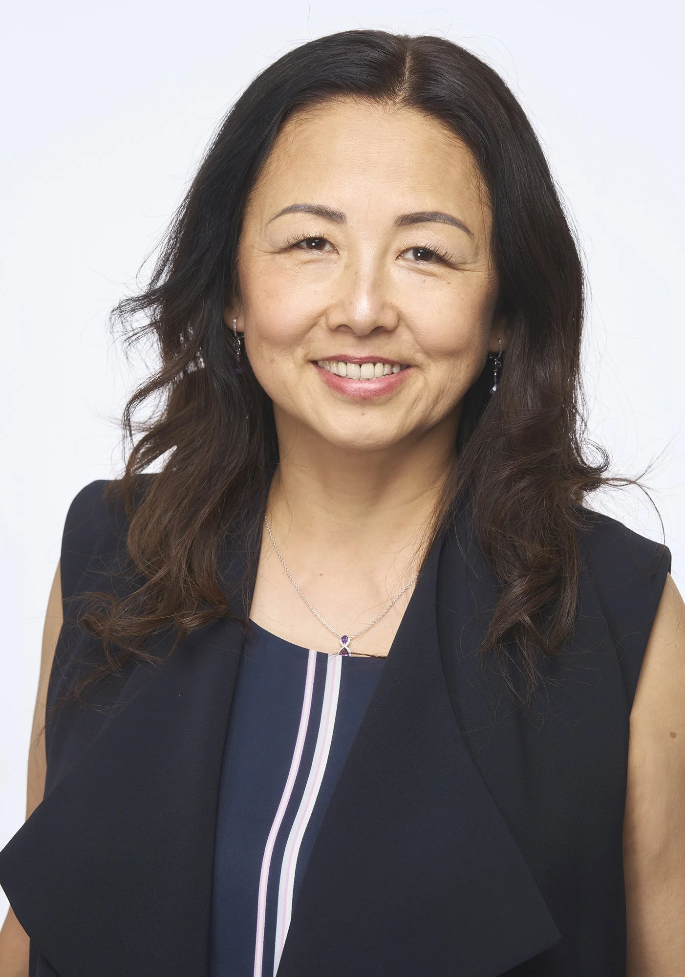Headshot of a smiling Asian woman with dark wavy hair, wearing a black sleeveless blazer, a navy top with a vertical stripe, and jewelry including earrings and a necklace, against a plain light background.