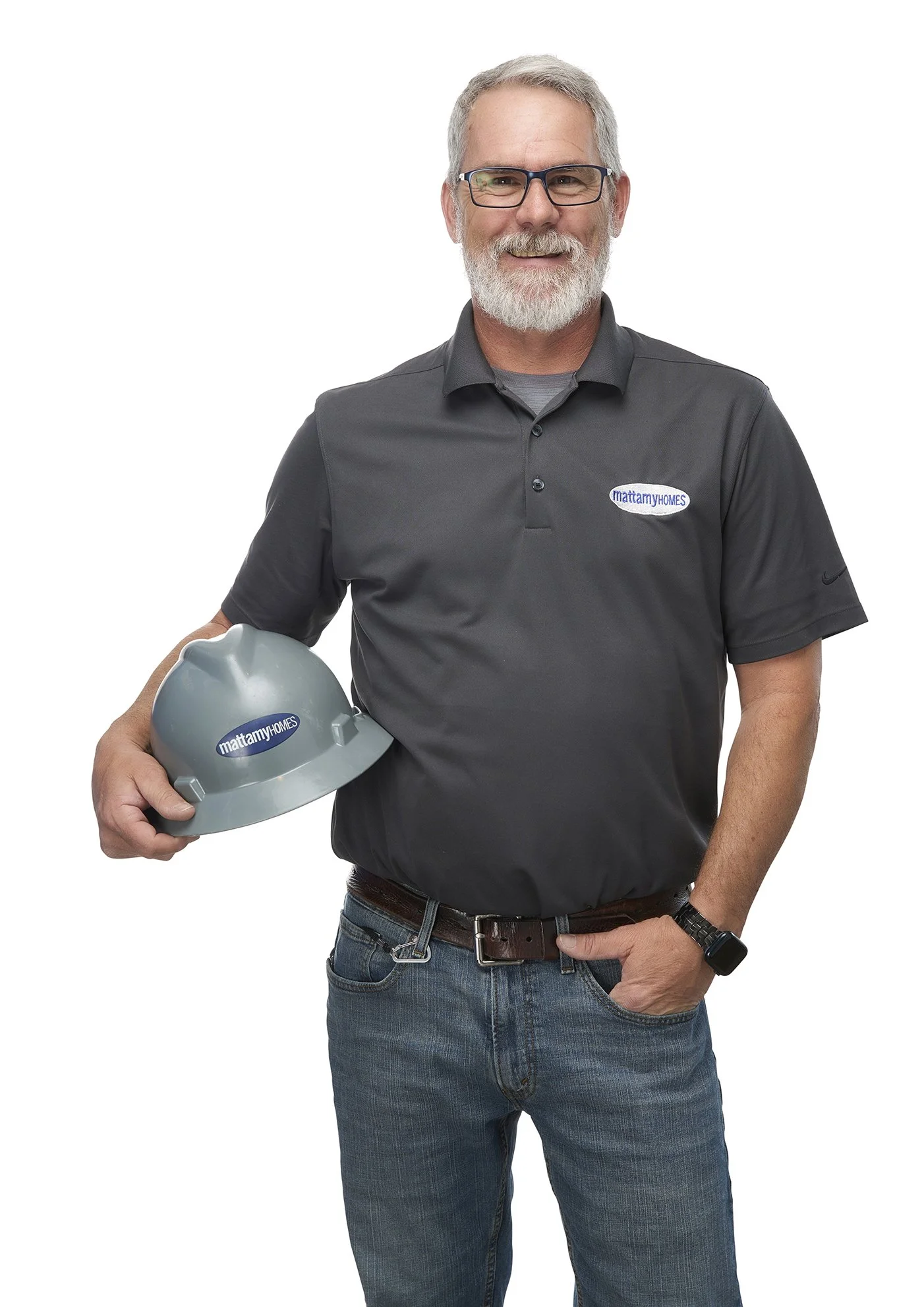 A smiling man with gray hair, beard, and glasses holding a gray construction helmet with 'mattamyHOMES' logo, wearing a gray polo shirt with the same logo, blue jeans, and a black smartwatch.