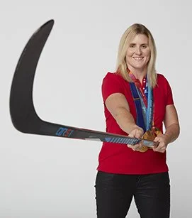 Woman in red shirt holding a hockey stick in front of a plain background.
