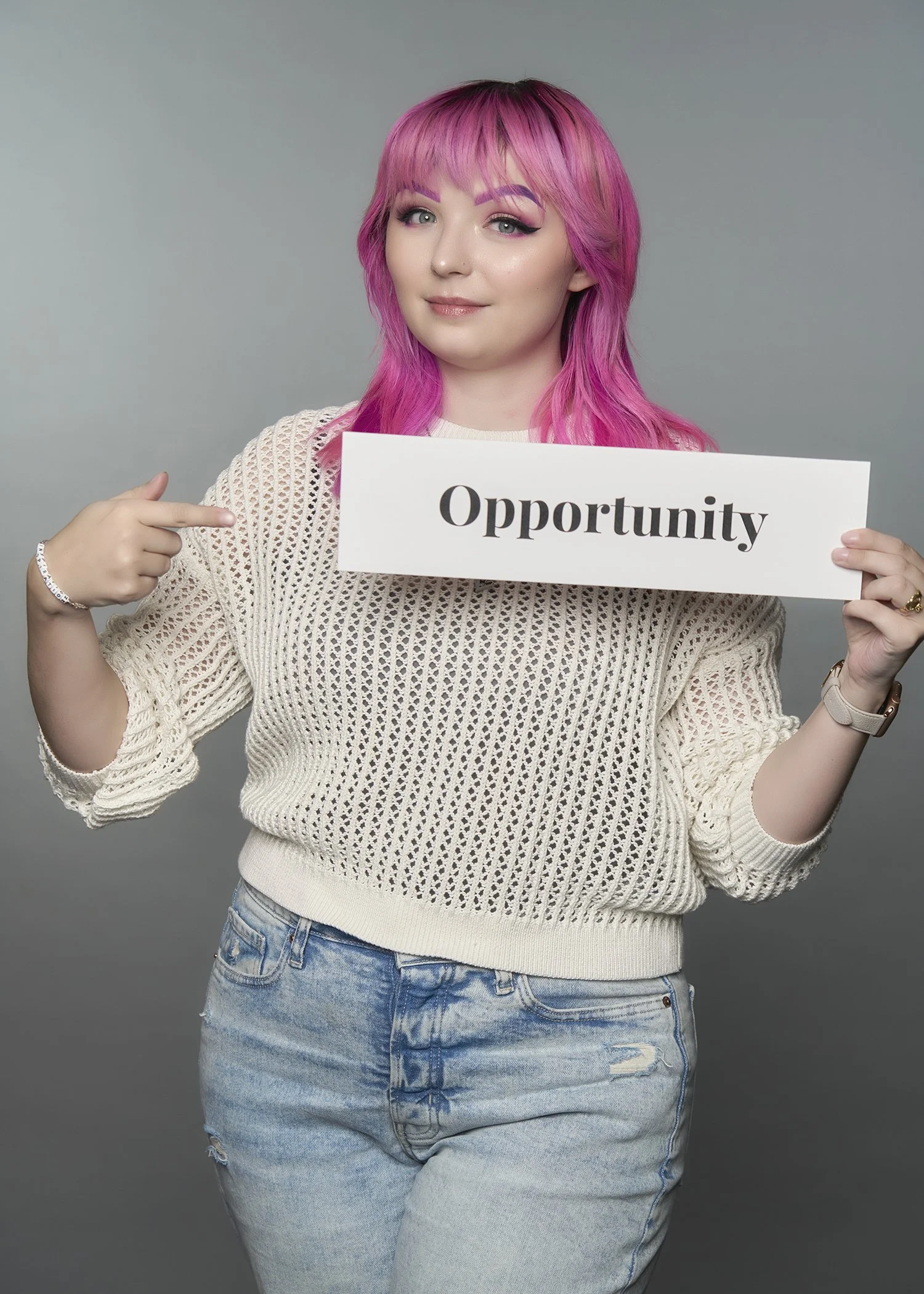 A young woman with pink hair and makeup, wearing a knit sweater and jeans, pointing to a sign that says 'Opportunity'.