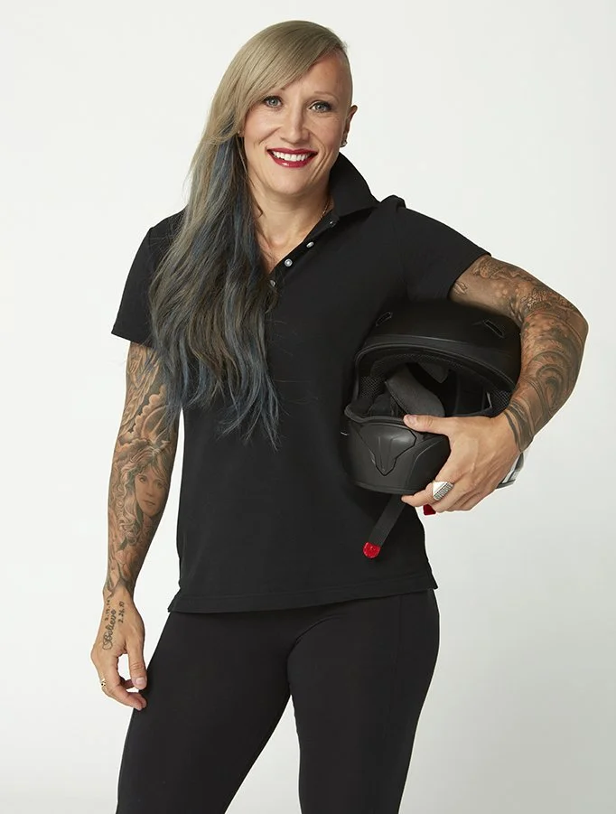 A smiling woman with long, multi-colored hair tattoos on her left arm, wearing a black polo shirt and black pants, holding a black motorcycle helmet against her hip, standing against a white background.