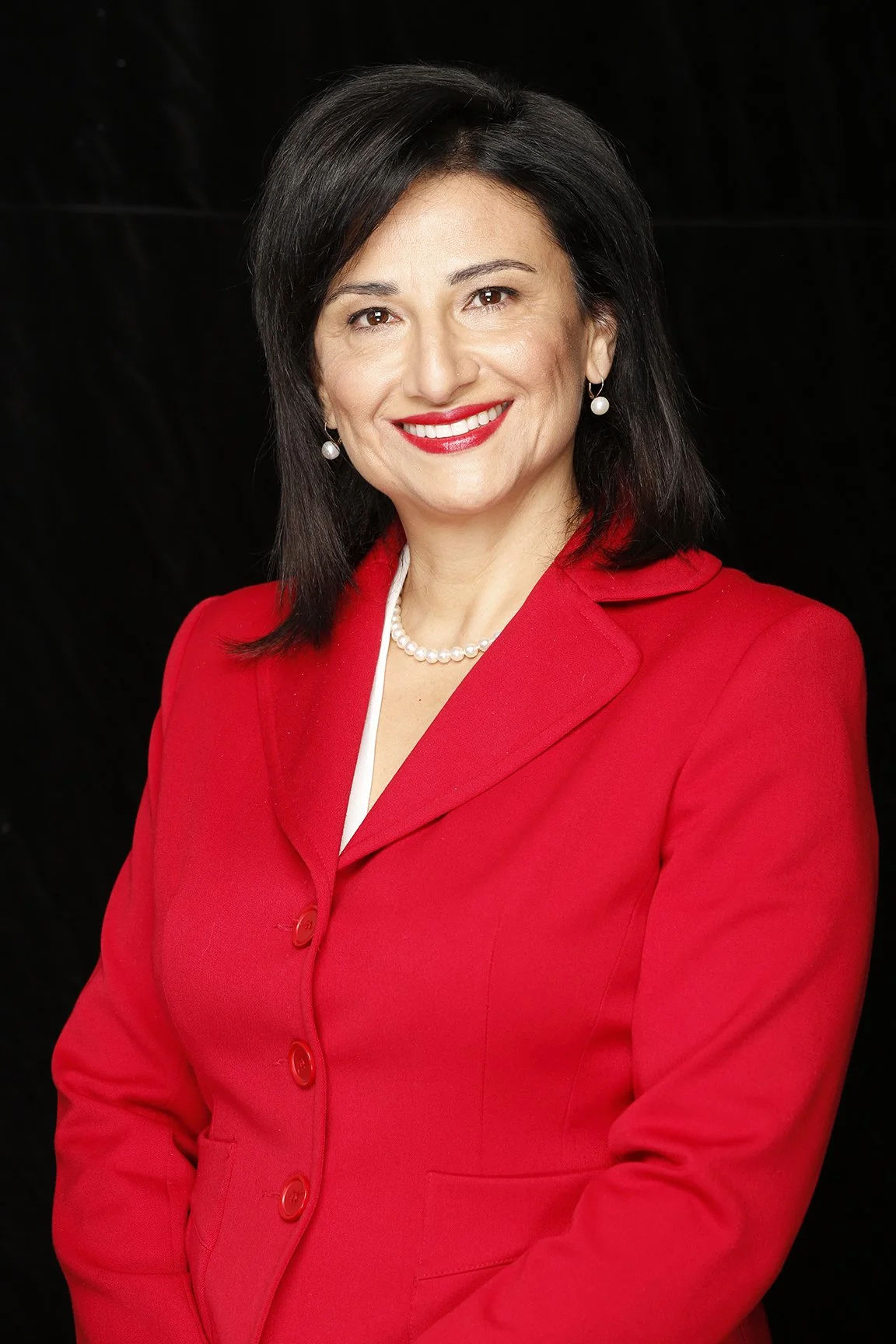 Portrait of a woman with dark hair, wearing a red blazer, pearl earrings, and a pearl necklace, smiling against a black background.