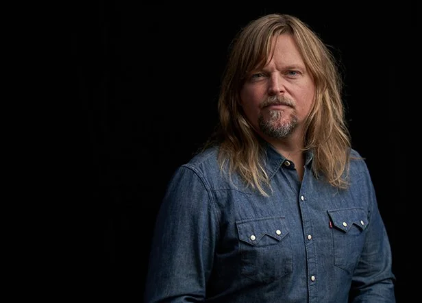 A man with long blonde hair, a beard, and mustache wearing a blue denim shirt against a black background.