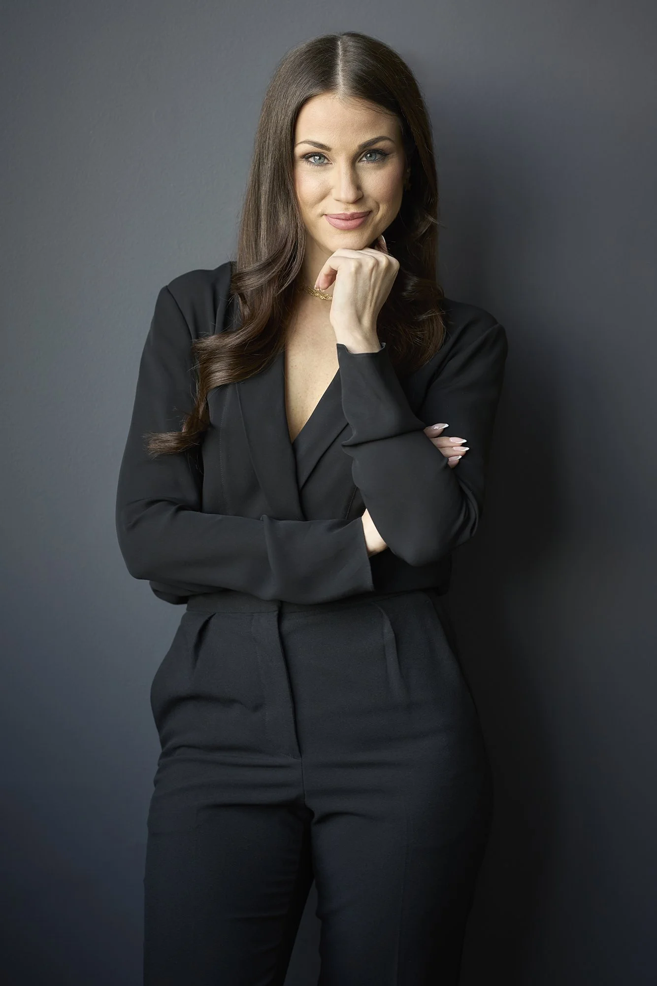 A woman with long brown hair in business attire, standing against a dark gray wall, looking at the camera with a confident expression.