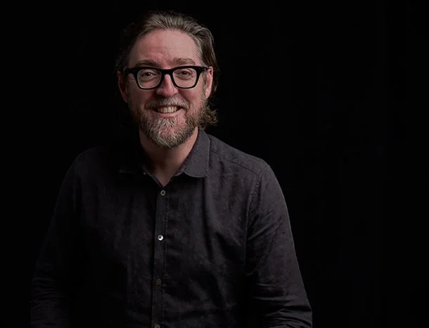A smiling man with glasses and a beard, wearing a dark button-up shirt, against a black background.