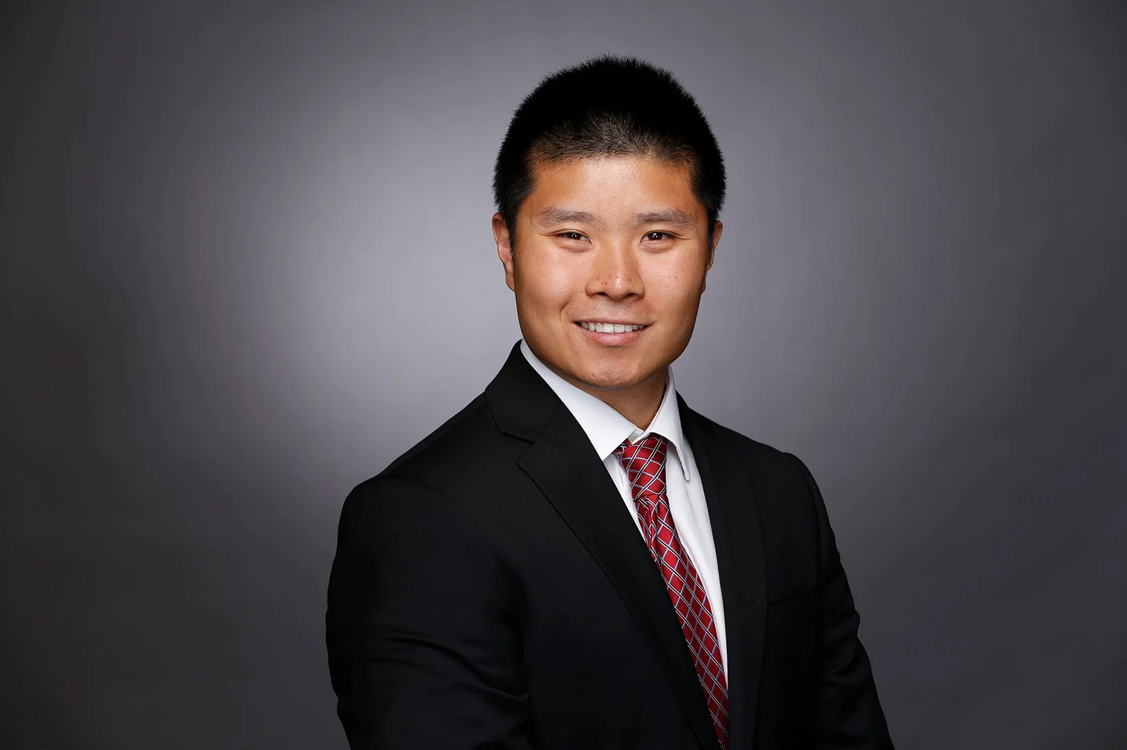 A young man wearing a black suit, white shirt, and red patterned tie, smiling in front of a dark gray gradient background.