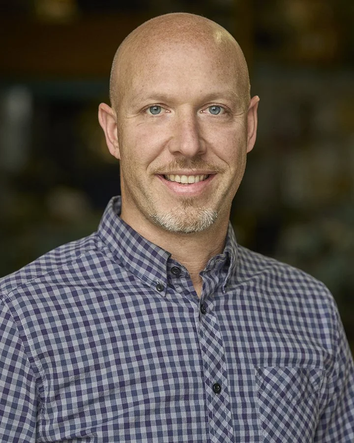 Headshot of a bald man with blue eyes, smiling, wearing a blue and gray checkered button-up shirt.
