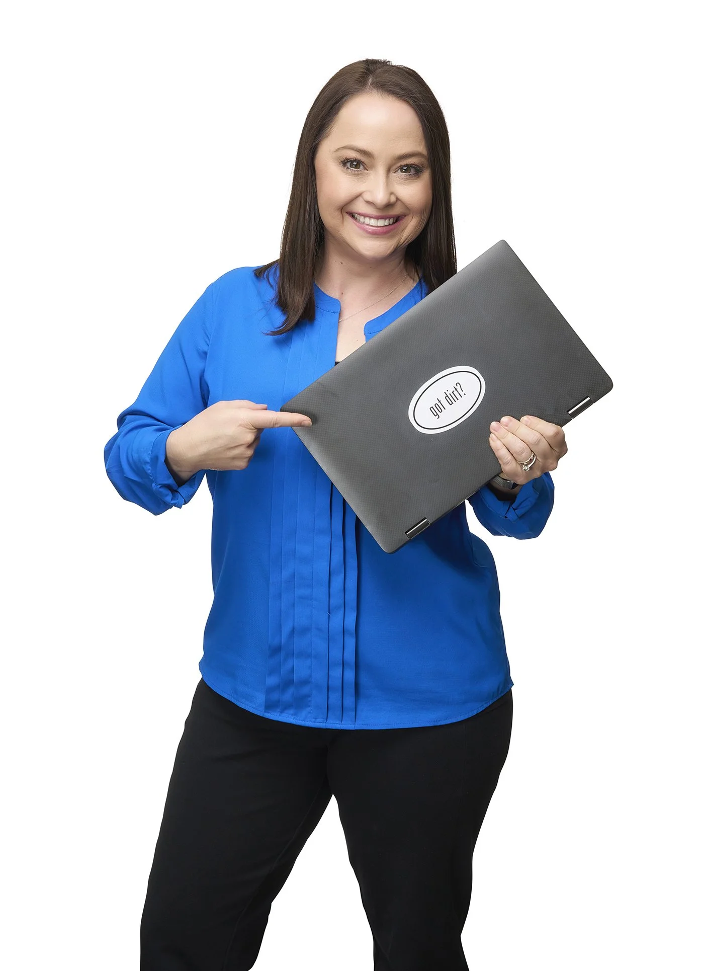 A woman with shoulder-length brown hair, wearing a blue blouse and black pants, is smiling and pointing at a closed laptop with a sticker that says "got dirt?" on a white background.
