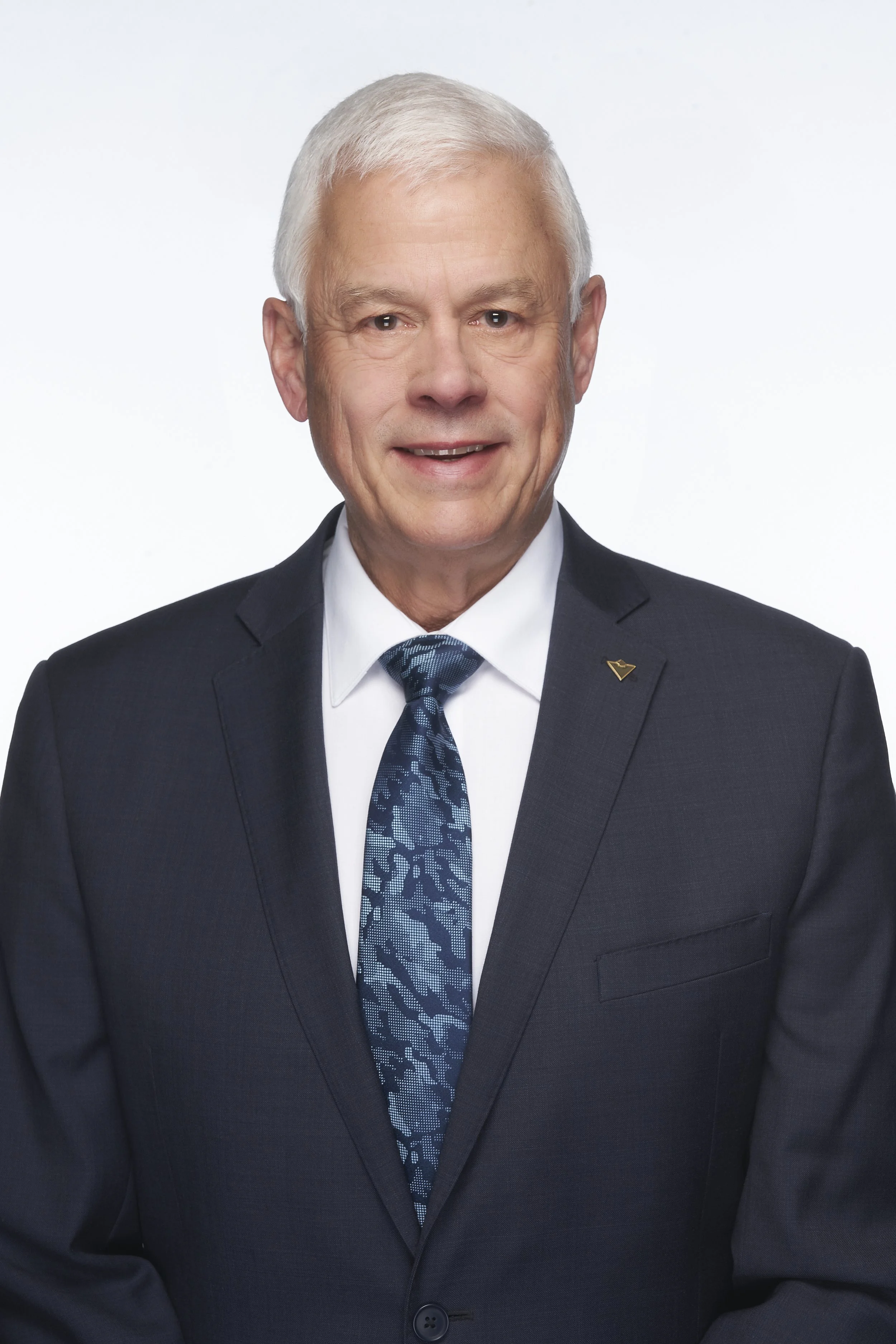 A senior man in a dark suit with a white shirt and a patterned blue tie, smiling at the camera against a plain background.
