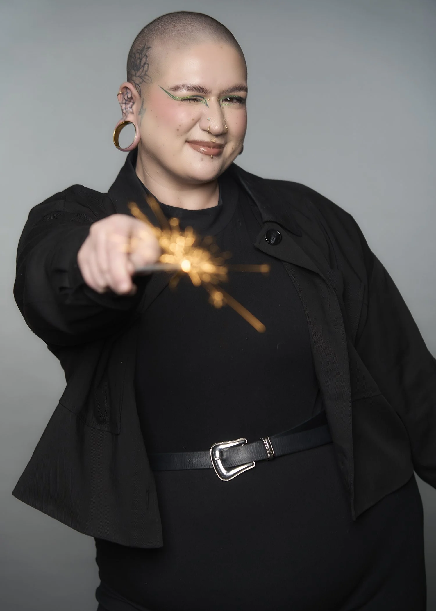 Person with a shaved head and tattoos, winking and smiling, holding a lit sparkler, wearing a black outfit and large earrings, against a plain grey background.