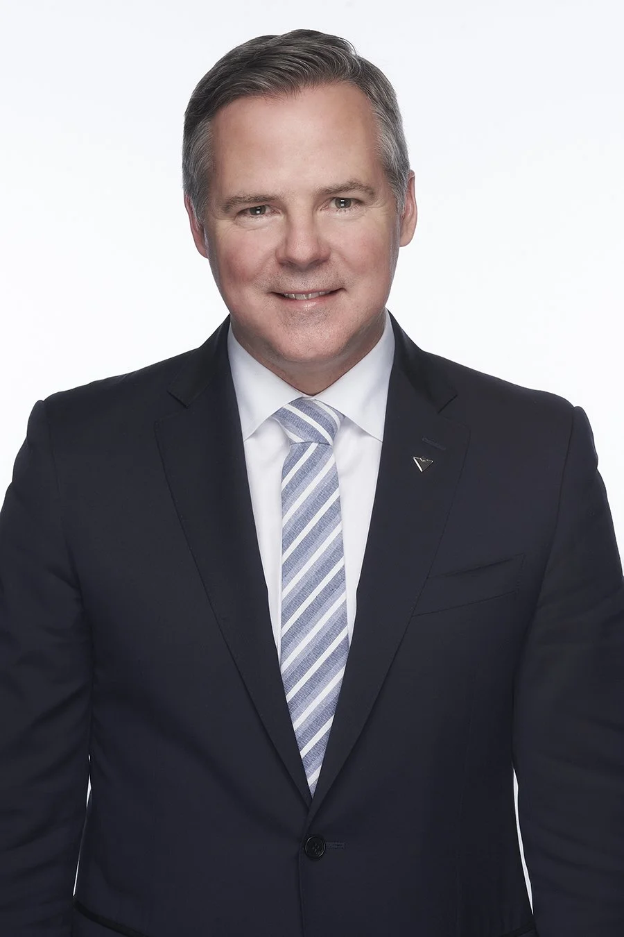 A professional man wearing a black suit, white dress shirt, and a striped tie, smiling at the camera against a plain light background.