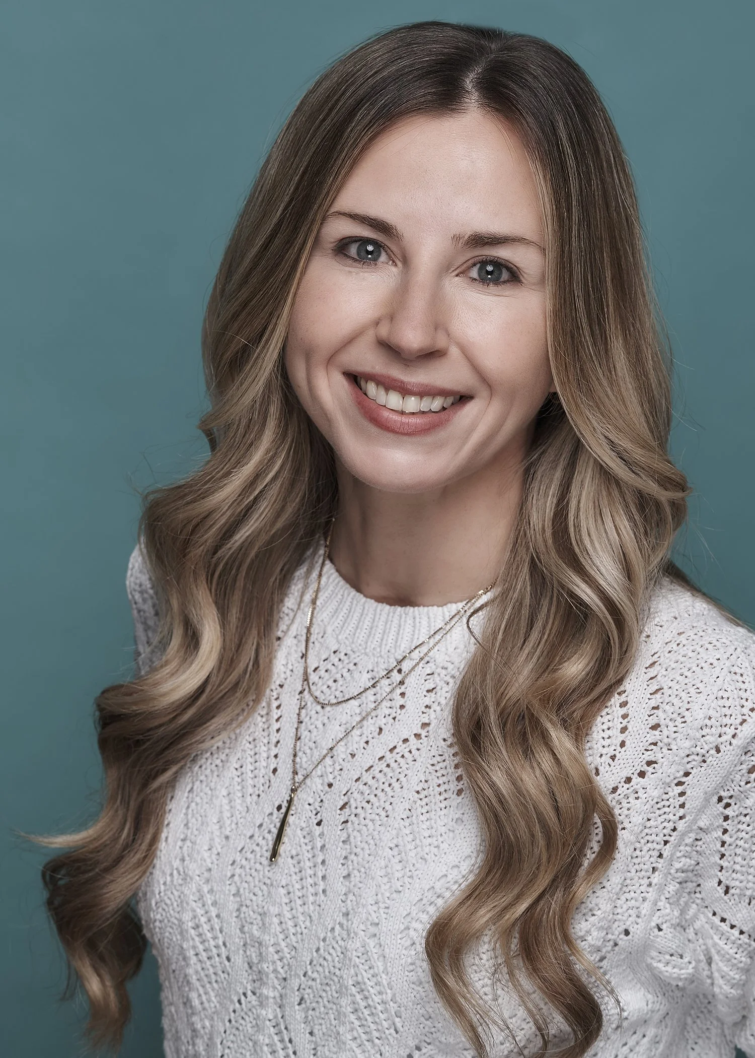 A woman with long, wavy blonde hair smiling against a teal background, wearing a white knit sweater and layered gold necklaces.