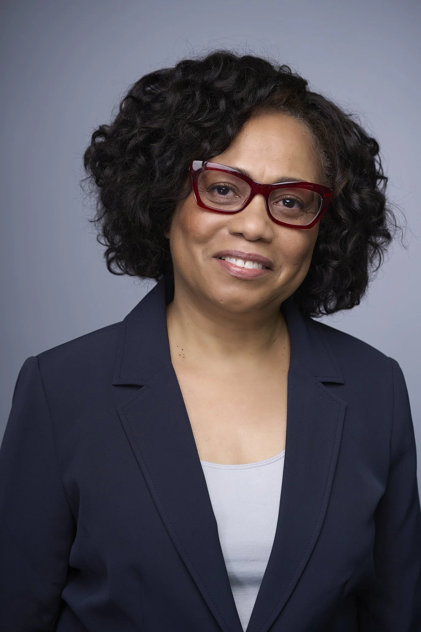 A professional woman with curly black hair and red glasses posing in front of a gray backdrop.