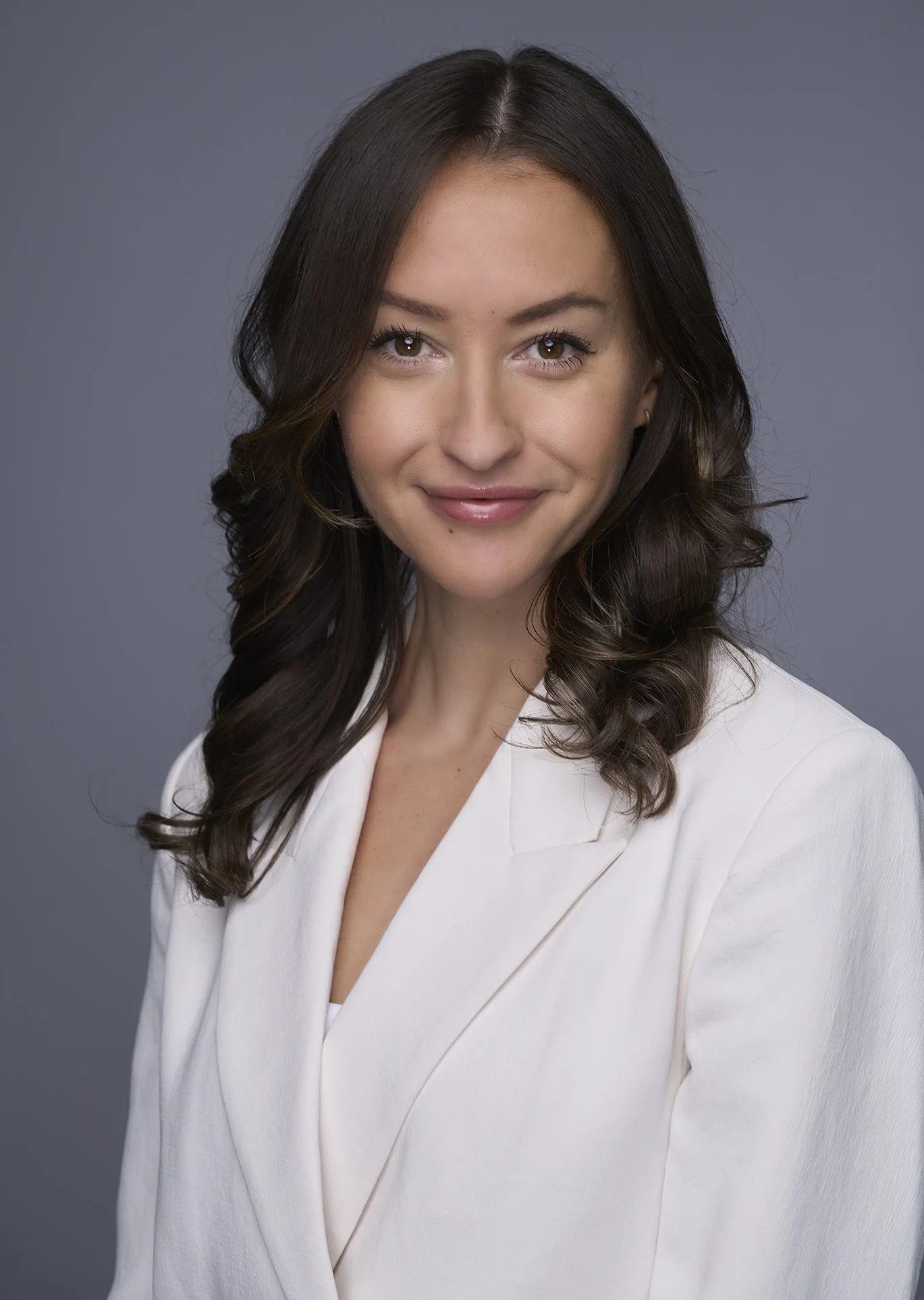 A woman with brown, wavy hair and brown eyes wearing a white blazer smiling at the camera against a gray background.